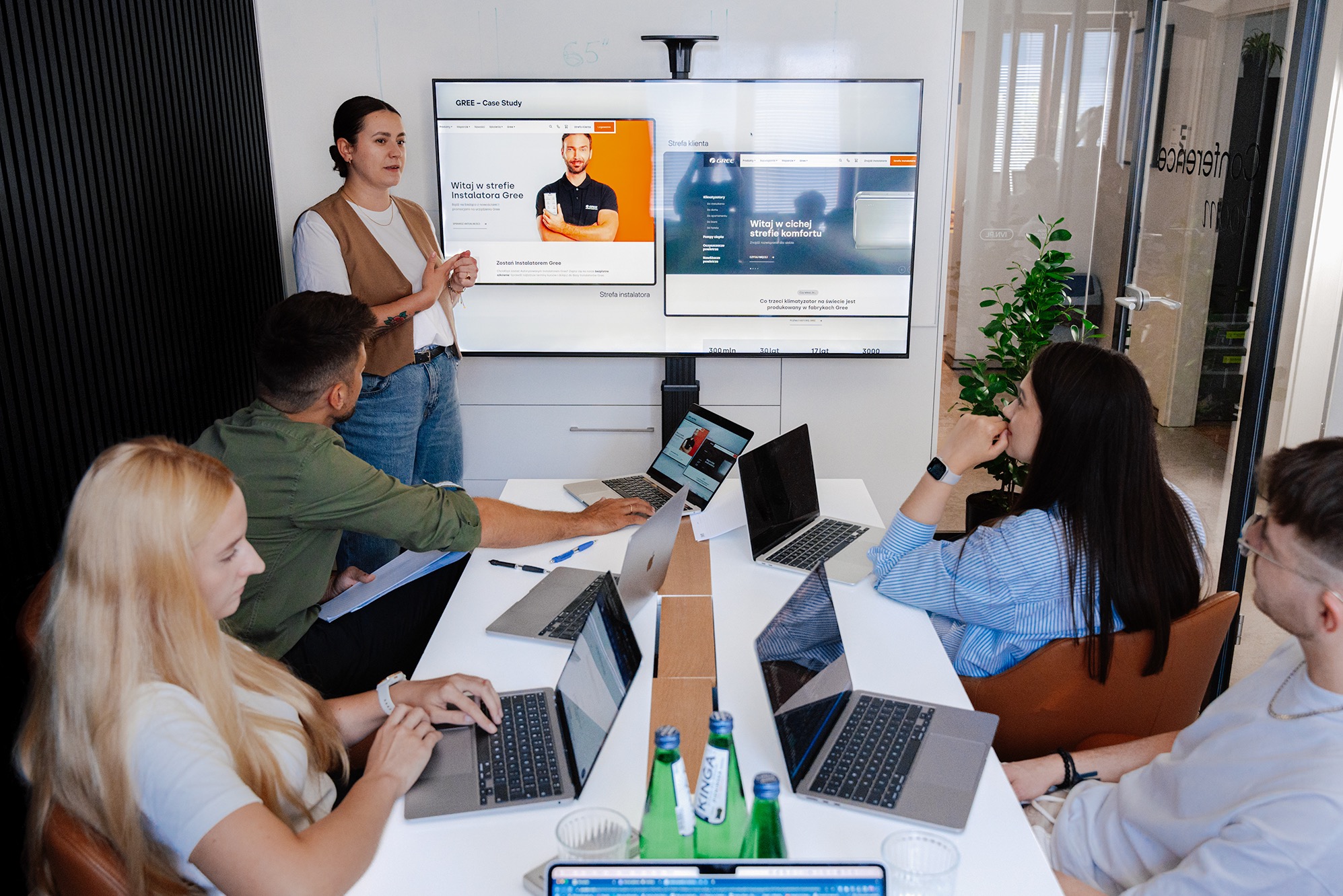 Prezentacja case study Gree: kobieta omawia stronę www na ekranie, zespół pracuje na laptopach przy stole. Jasne biuro, butelki wody na stole.