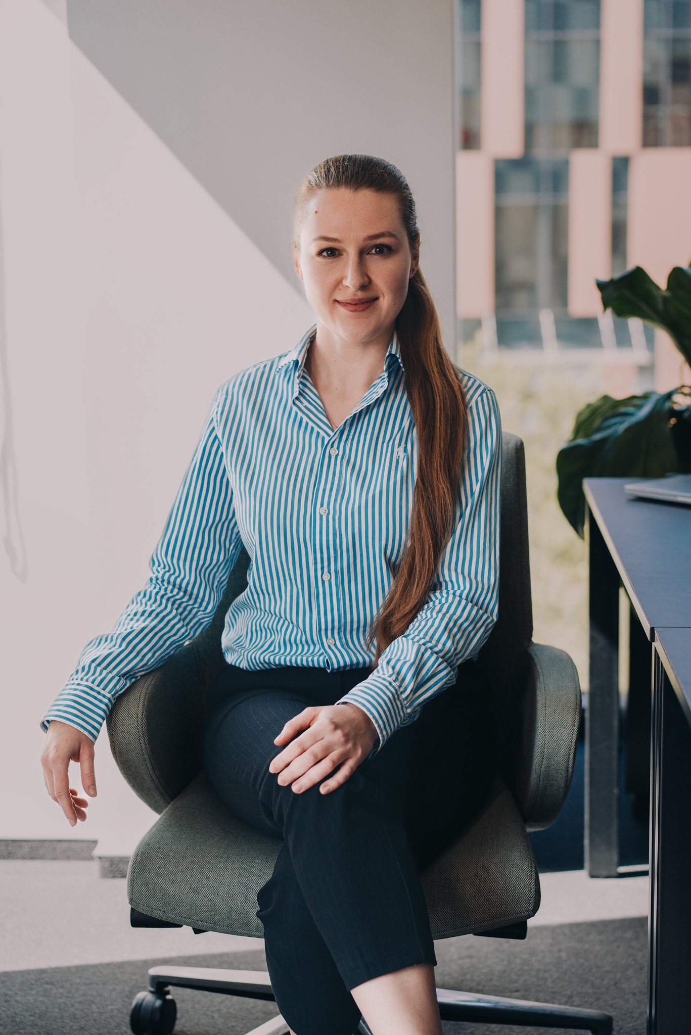 Portret kobiety w błękitnej koszuli w paski, siedzącej na krześle w biurze. Uśmiechnięta, z długimi włosami. W tle okno i roślina doniczkowa.