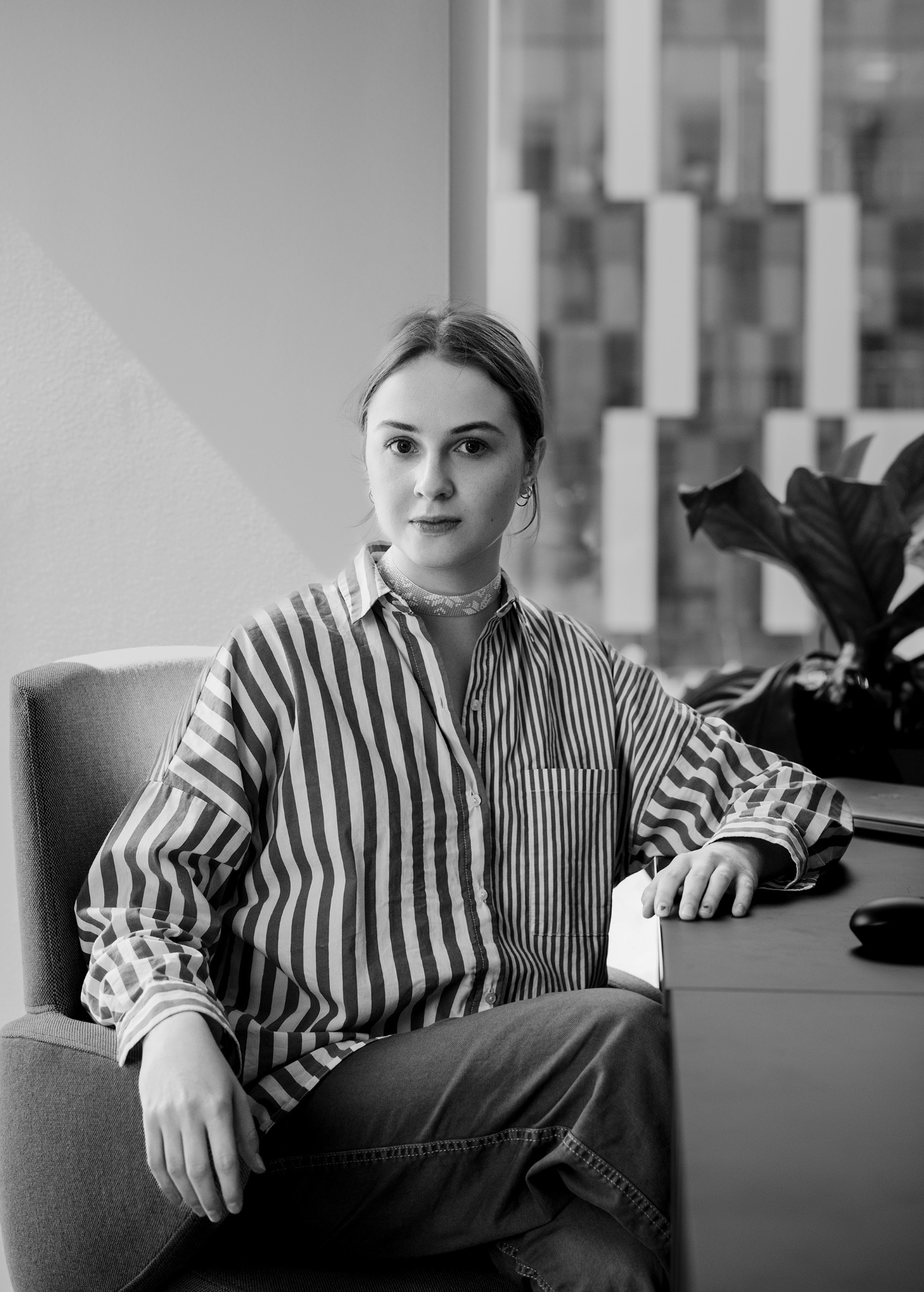 Portret kobiety w paski koszuli, siedzącej przy biurku w nowoczesnym wnętrzu. Stonowana kolorystyka, naturalne światło, minimalistyczne tło z rośliną.