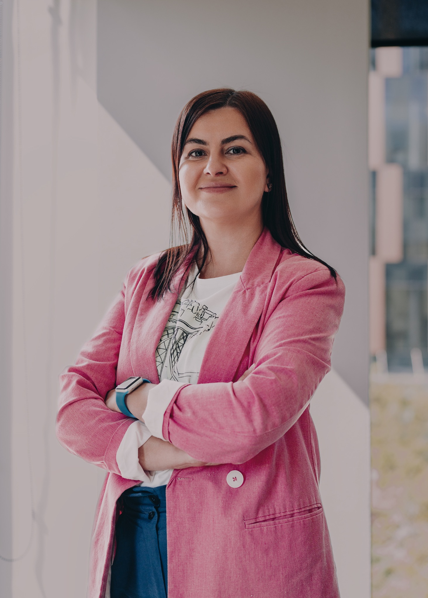 Portret kobiety w różowym żakiecie, z założonymi rękami, na tle jasnego wnętrza z widocznym fragmentem budynku za oknem. Uśmiechnięta, pewna siebie.