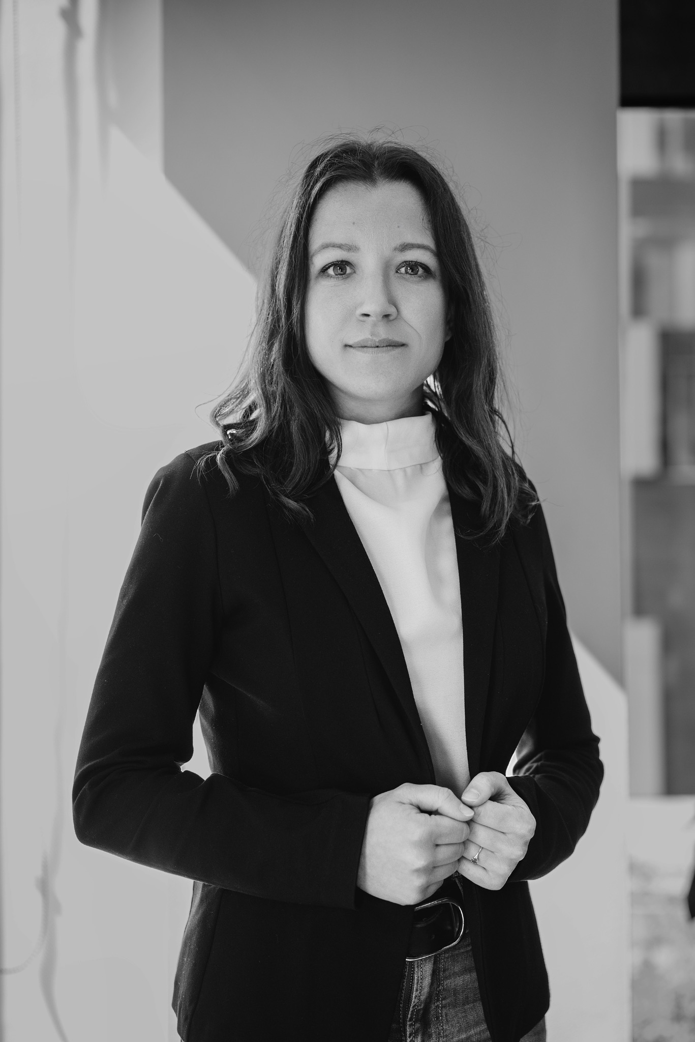 Czarno-biały portret kobiety w marynarce, z delikatnym uśmiechem, na neutralnym tle. Dłonie splecione przy zapięciu marynarki, subtelne światło podkreśla rysy twarzy.