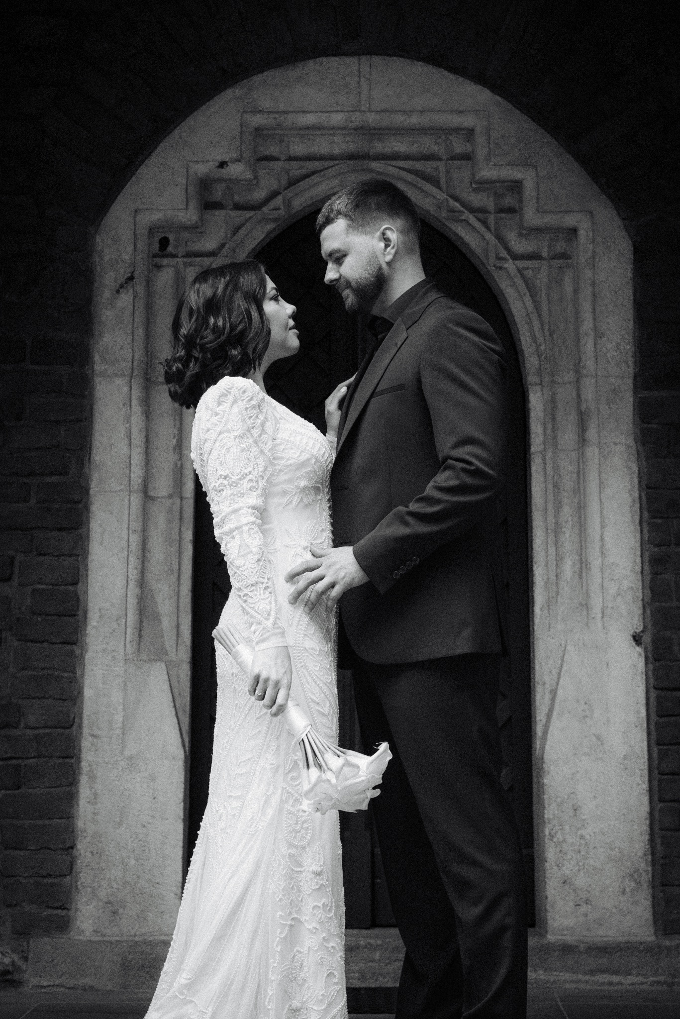 Czarno-białe zdjęcie pary młodej w eleganckich strojach, stojącej w kamiennym portalu. Panna młoda trzyma bukiet. Ujęcie z dołu podkreśla architekturę.