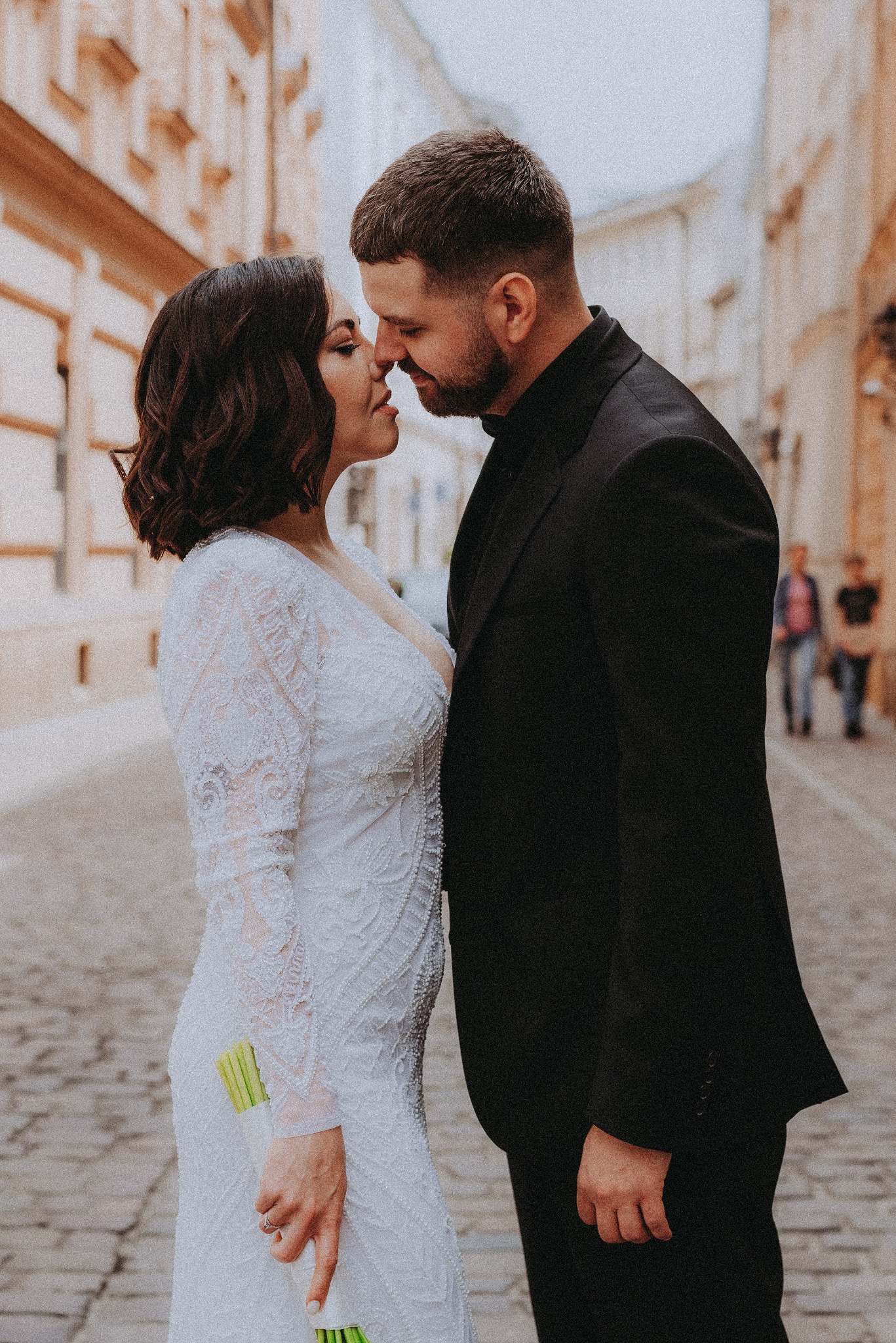 Para młoda w zbliżeniu, niemal dotykająca się nosami na tle starego miasta. Panna młoda w koronkowej sukni, pan młody w czarnym garniturze. Romantyczna atmosfera.