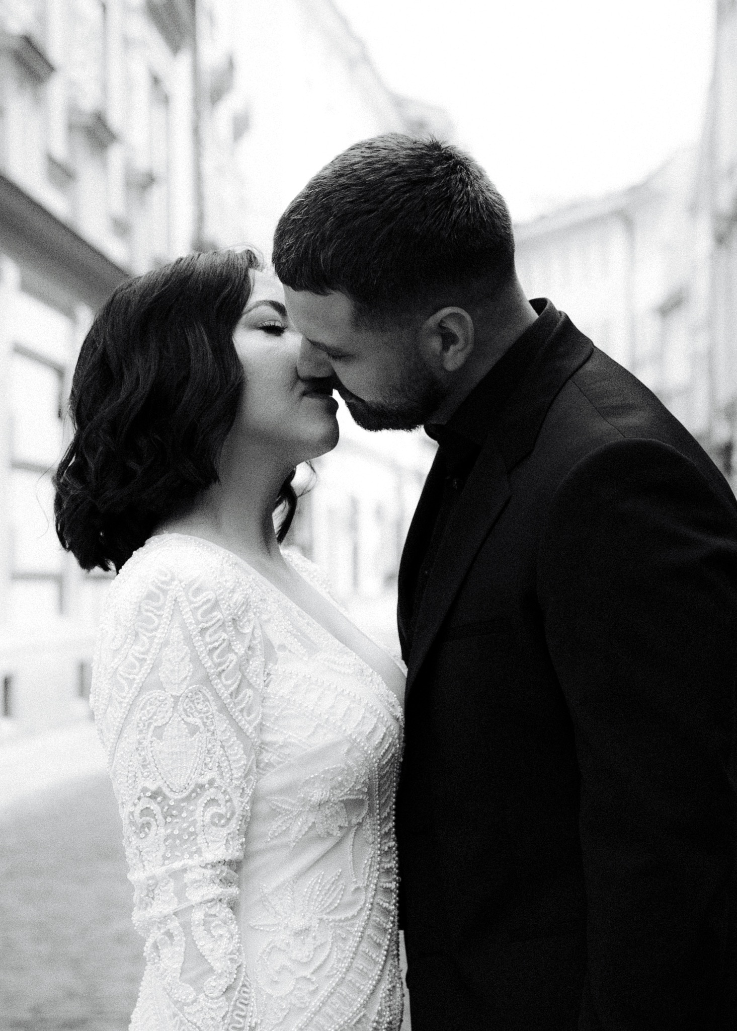 Czarno-białe zbliżenie całującej się pary młodej na tle miejskiej uliczki. Ona w koronkowej sukni, on w czarnym garniturze. Romantyczna fotografia ślubna.