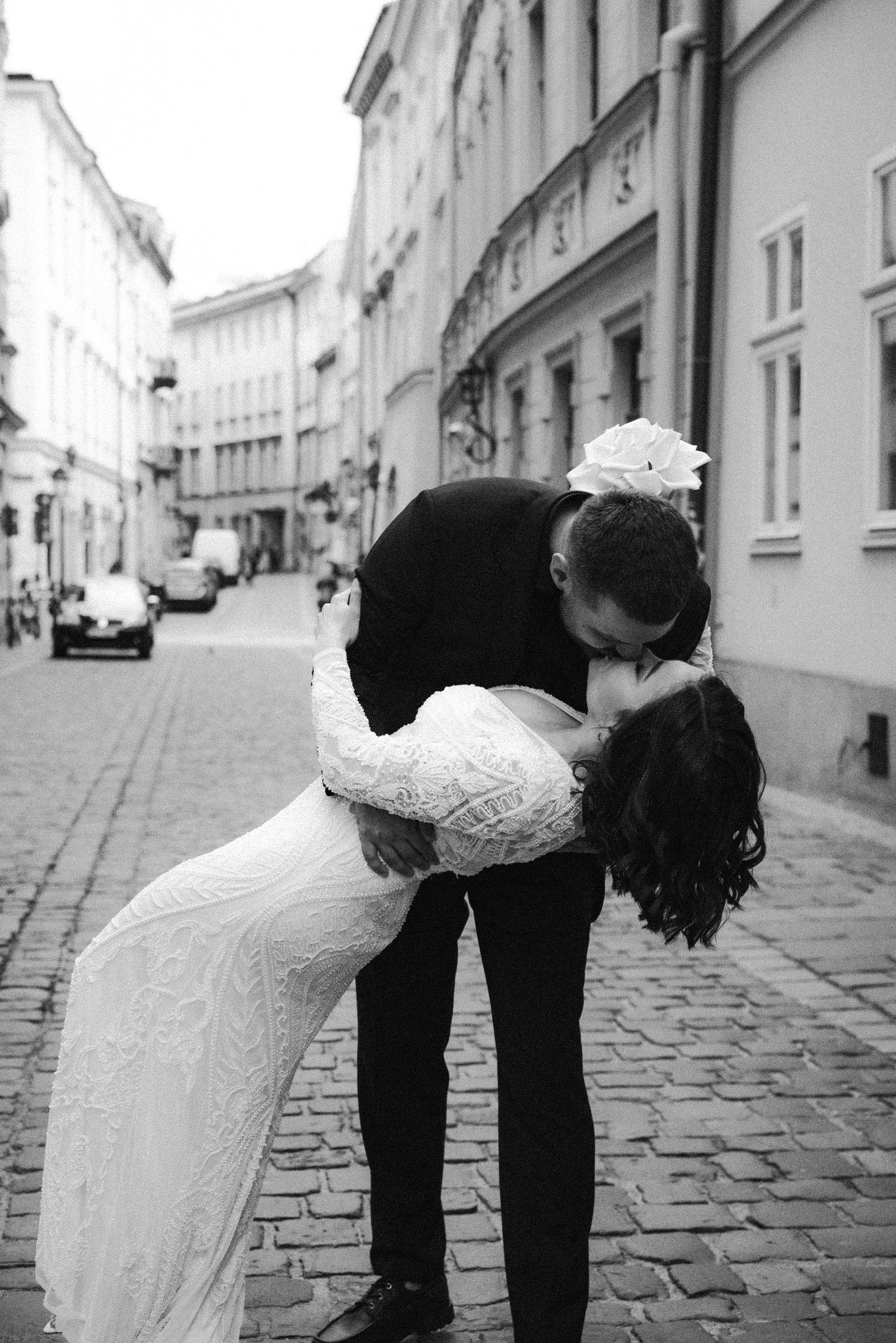 Czarno-białe zdjęcie: para młoda w pocałunku na brukowanej ulicy, panna młoda w koronkowej sukni, pan młody w garniturze, w tle budynki miejskie.