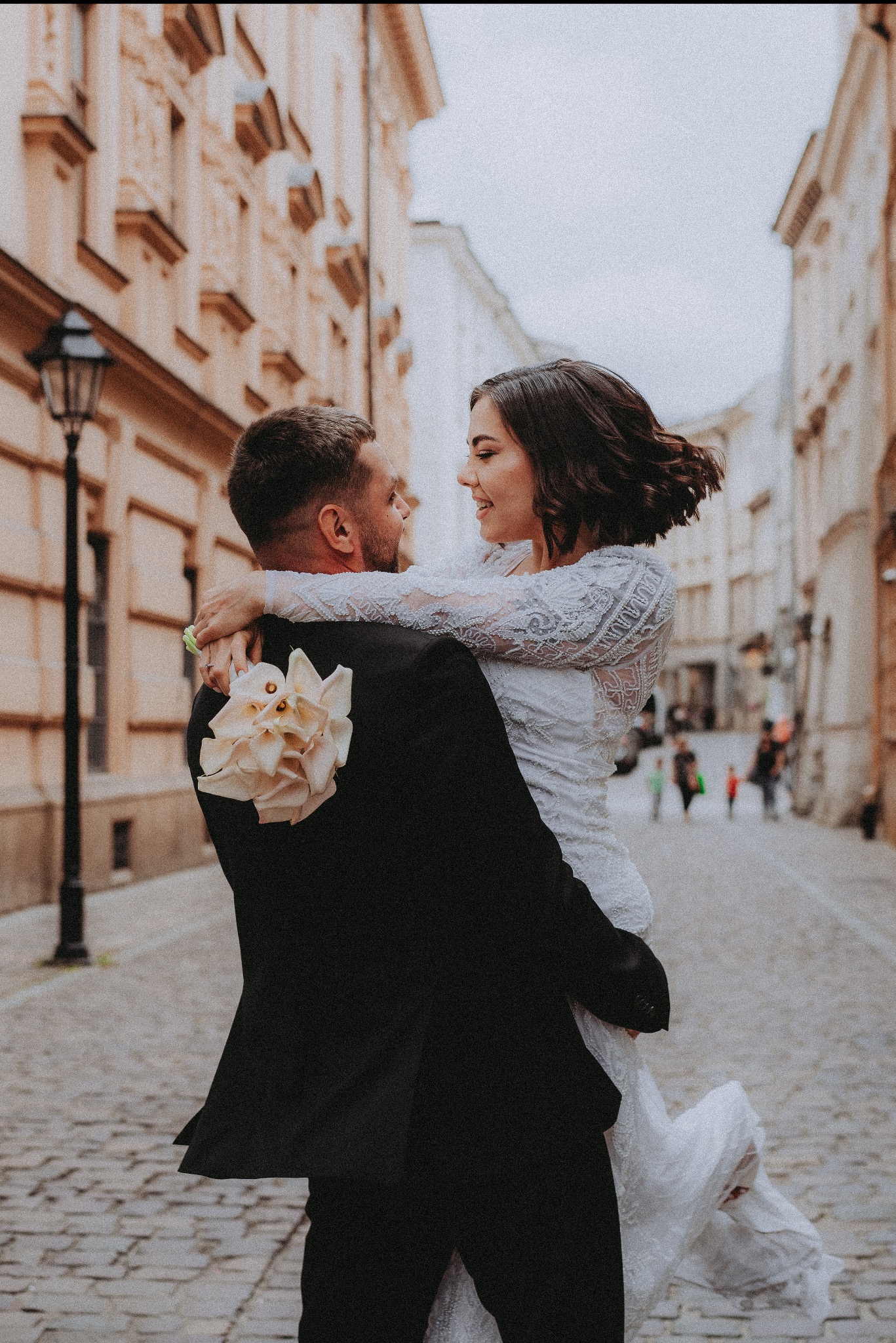 Uśmiechnięta panna młoda w białej sukni na rękach pana młodego z bukietem na tle uliczki starego miasta. Romantyczna sesja ślubna w plenerze.