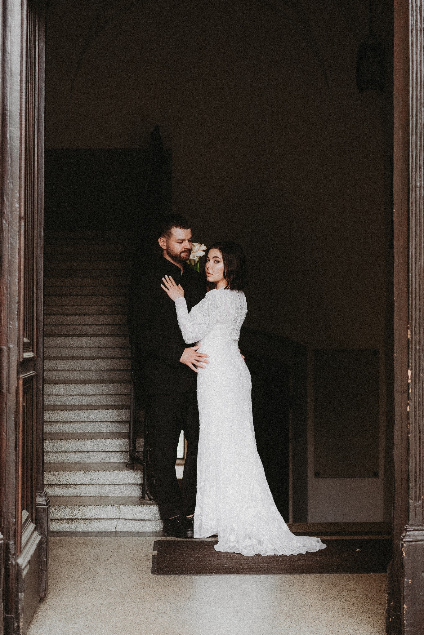 Elegancka para młoda w wejściu do budynku. Panna młoda w koronkowej sukni, pan młody w czarnym garniturze. Schody w tle. Ujęcie z dołu, podkreślające wysokość.