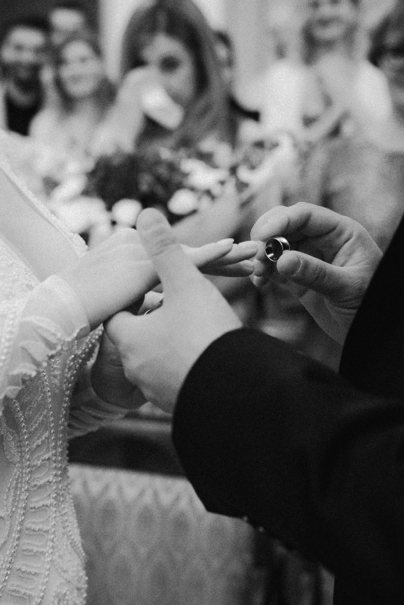 Czarno-białe zbliżenie dłoni nakładających obrączkę ślubną, w tle rozmyte twarze gości. Delikatna koronka sukni panny młodej.