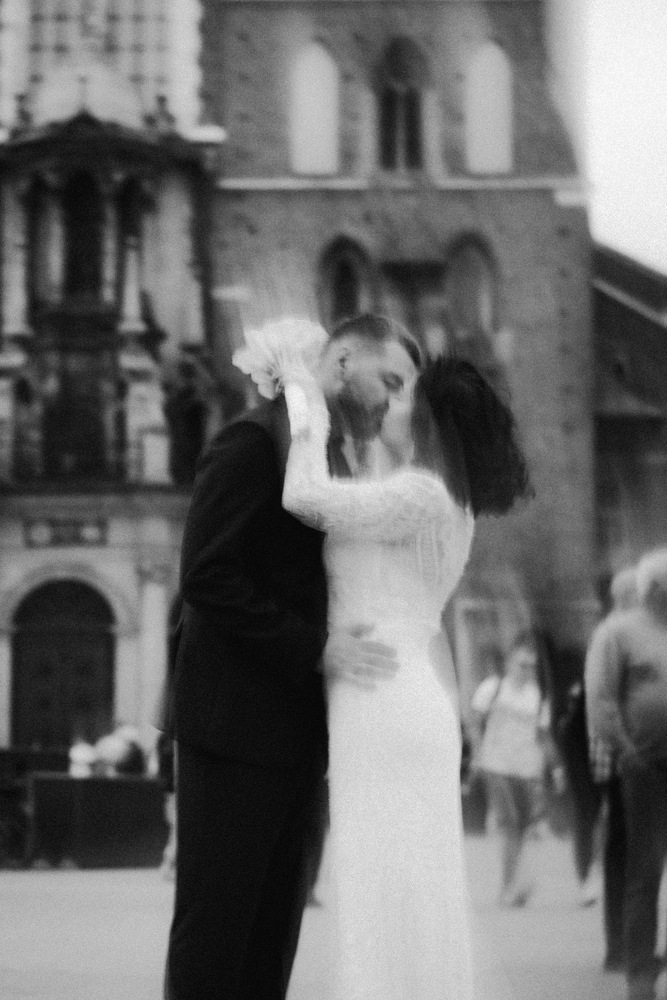 Czarno-białe, ziarniste zdjęcie całującej się pary młodej na tle zabytkowej architektury. Panna młoda w koronkowej sukni, pan młody w ciemnym garniturze. Rozmycie dodaje intymności.