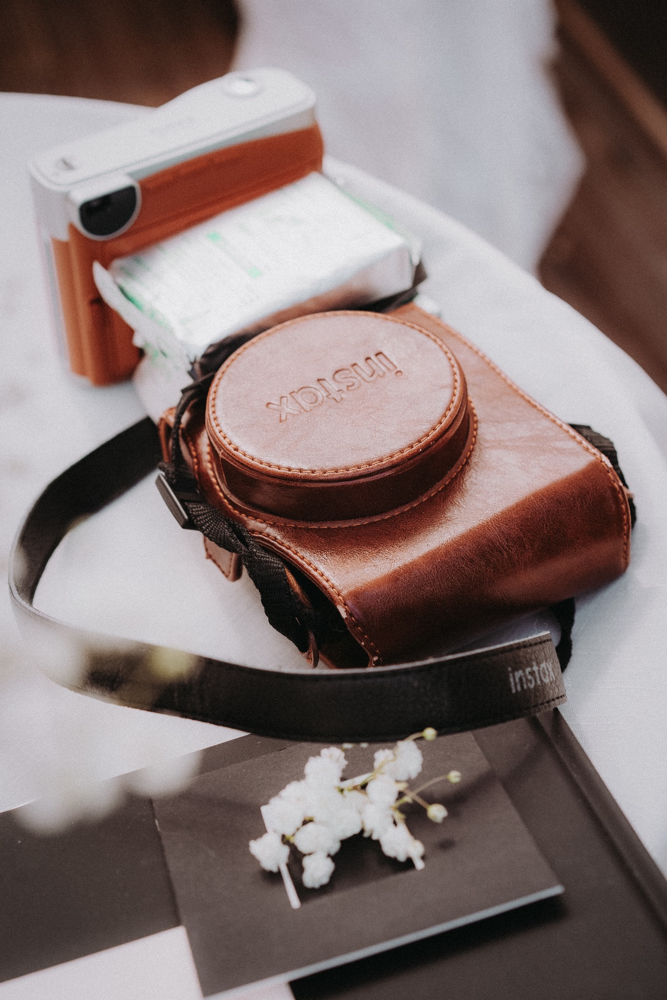 Brązowy futerał na aparat Instax z paskiem, aparat i kwiaty na czarno-białym tle. Delikatne światło podkreśla detale.