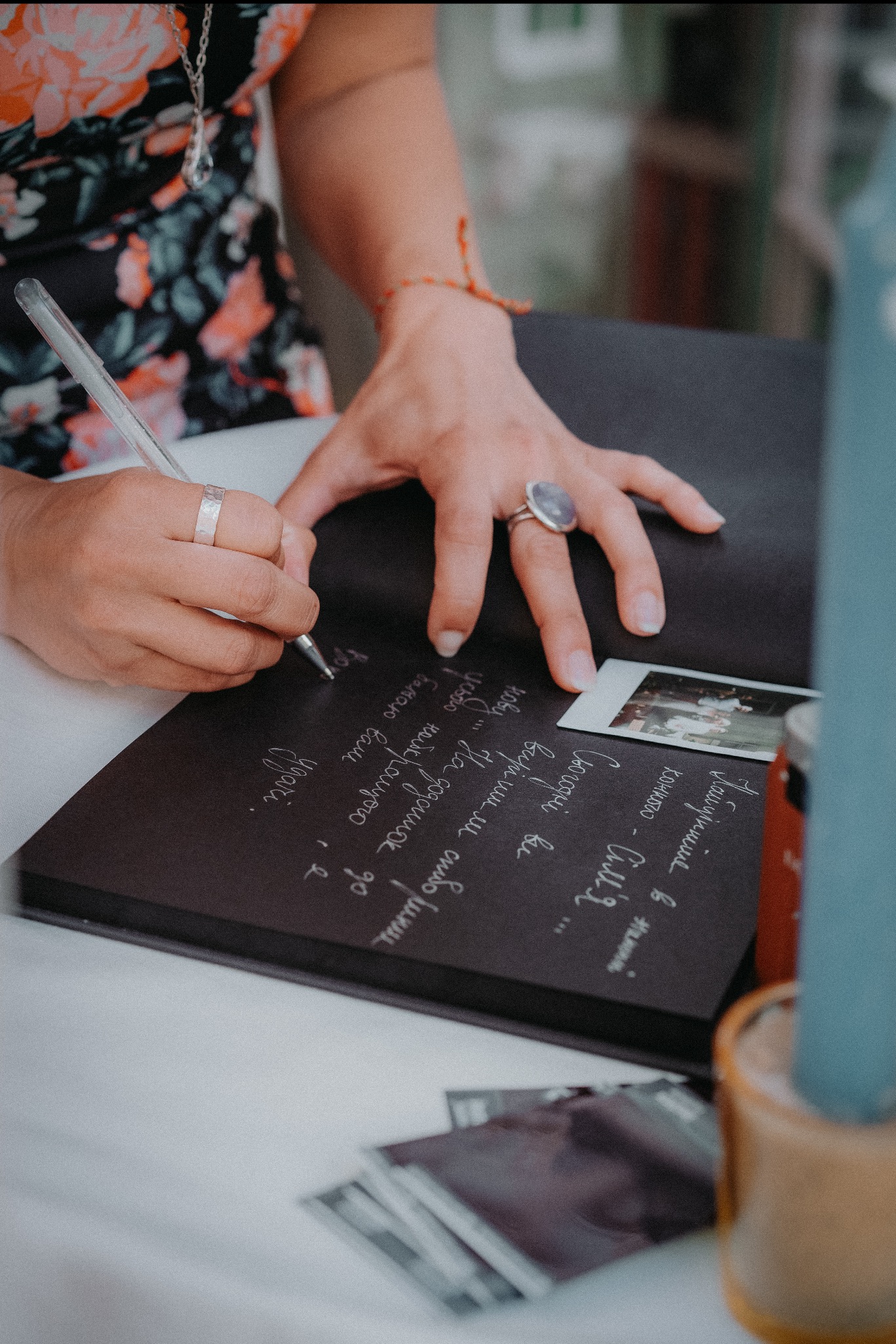Osoba wpisuje życzenia do czarnej księgi gości obok zdjęcia polaroidowego i stosu kart, w tle widać fragment dekoracji i osoby w kwiecistej sukience.