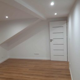 Kamil Domagała - Jasne wnętrze po remoncie: drewnopodobne panele, białe ściany i drzwi z poziomymi frezami. Oświetlenie punktowe w suficie.