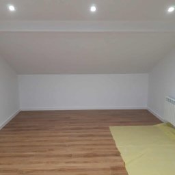 Kamil Domagała - Wnętrze po remoncie: świeżo pomalowane białe ściany, drewnopodobne panele podłogowe częściowo zakryte folią ochronną, widoczny grzejnik i oświetlenie sufitowe.