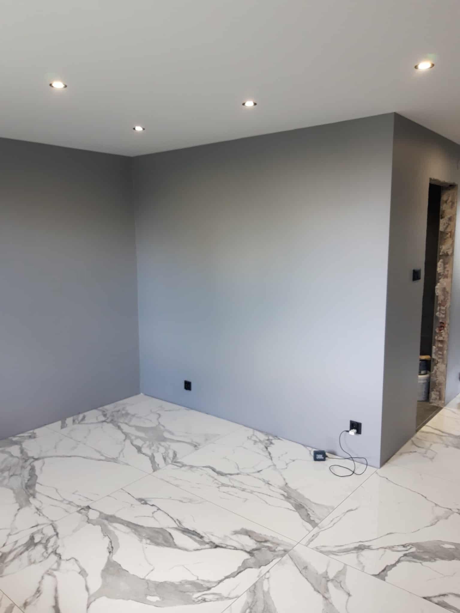 Pomieszczenie po remoncie: szare ściany, marmurowe płytki podłogowe. Widoczne gniazdka elektryczne i oświetlenie sufitowe. Fragment przejścia do innego pomieszczenia.