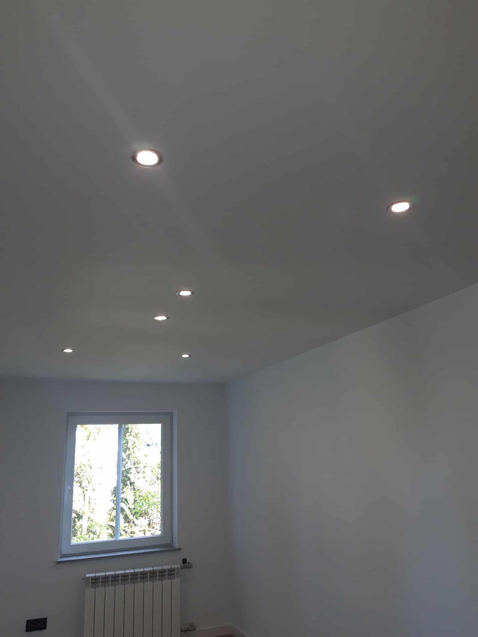 Białe wnętrze po remoncie: gładkie ściany, sufit z punktowym oświetleniem LED, okno z widokiem na zieleń i grzejnik pod nim. Minimalistyczny design.