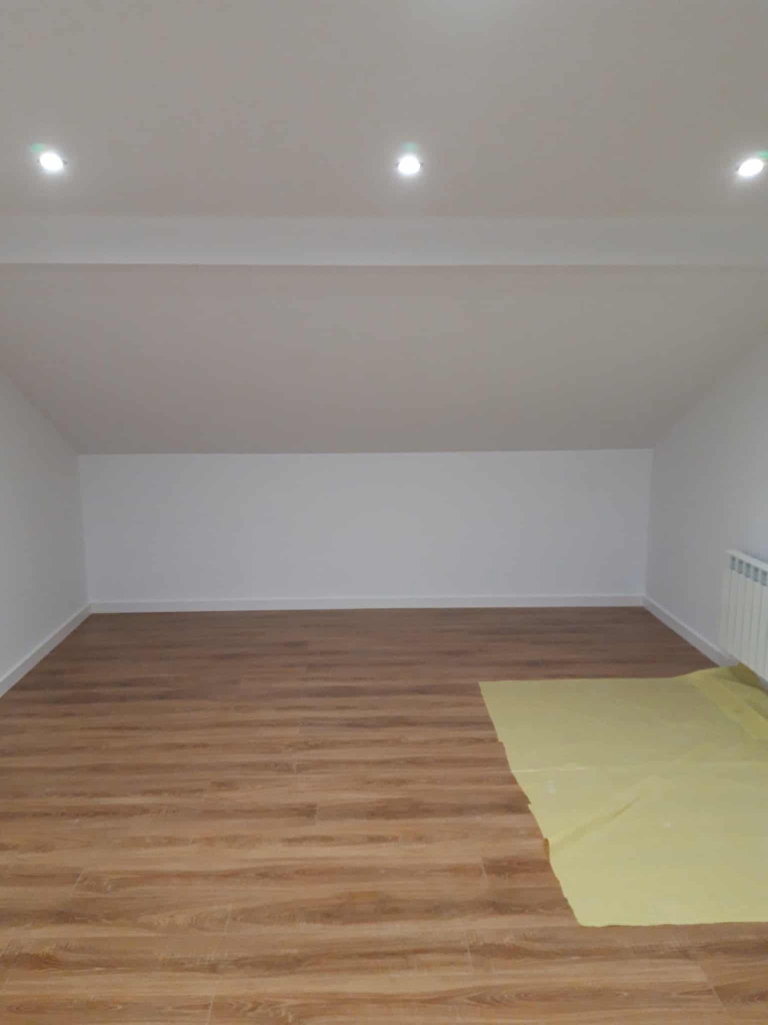 Wnętrze po remoncie: świeżo pomalowane białe ściany, drewnopodobne panele podłogowe częściowo zakryte folią ochronną, widoczny grzejnik i oświetlenie sufitowe.