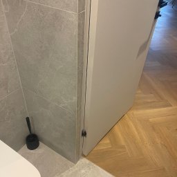 Oleksandr Potapchuk - Wykończona łazienka z szarą, kamienną okładziną ścian i podłogi, białą toaletą i otwartymi, białymi drzwiami, za którymi widać parkiet ułożony w jodełkę.