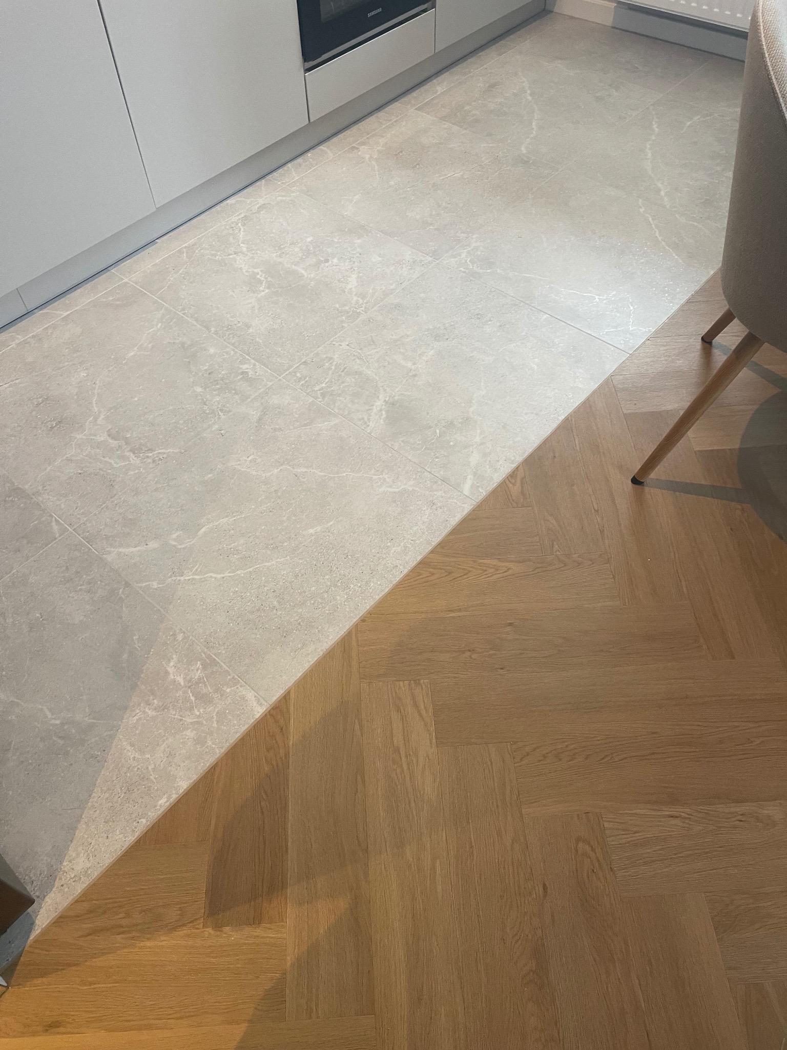 Połączenie płytek imitujących marmur i parkietu ułożonego w jodełkę francuską w kuchni. Widoczny fragment krzesła i zabudowy kuchennej.