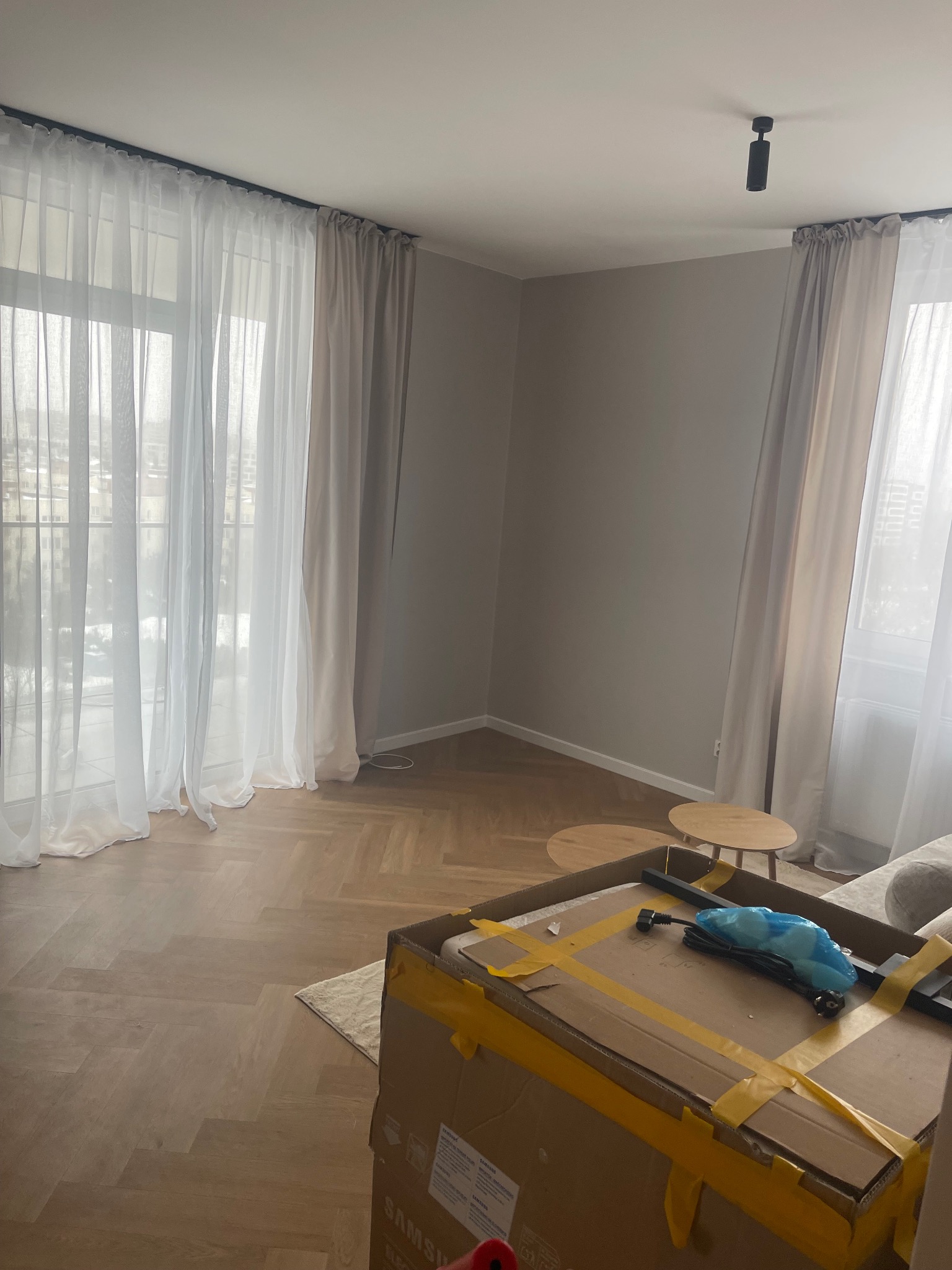 Jasne wnętrze z parkietem w jodełkę, okna z białymi zasłonami i karton z elementami oświetlenia. Widoczne małe stoliki kawowe i lampa sufitowa.