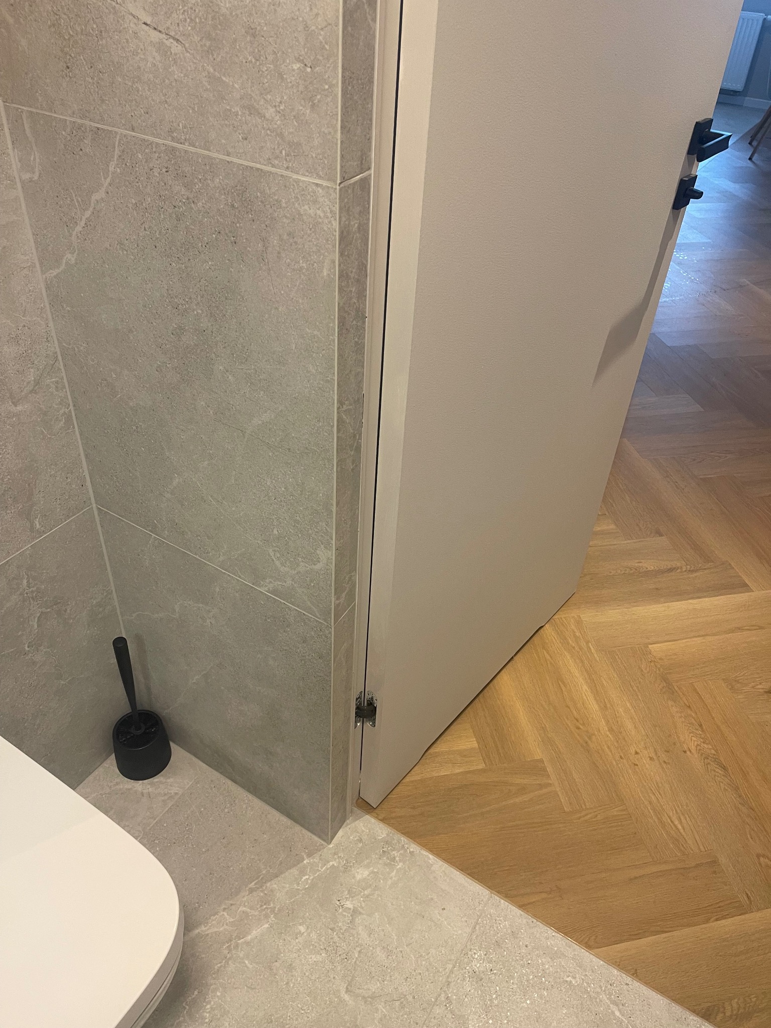 Wykończona łazienka z szarą, kamienną okładziną ścian i podłogi, białą toaletą i otwartymi, białymi drzwiami, za którymi widać parkiet ułożony w jodełkę.