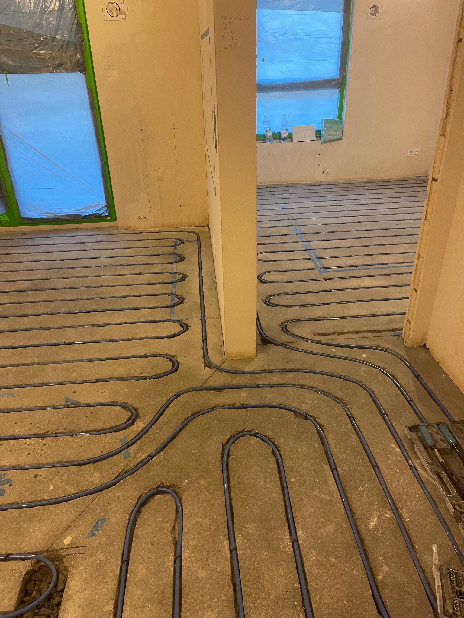 Niebieskie rury ogrzewania podłogowego ułożone na szarym betonie w trakcie prac remontowych. Okna zabezpieczone folią malarską. Widoczne narzędzia.