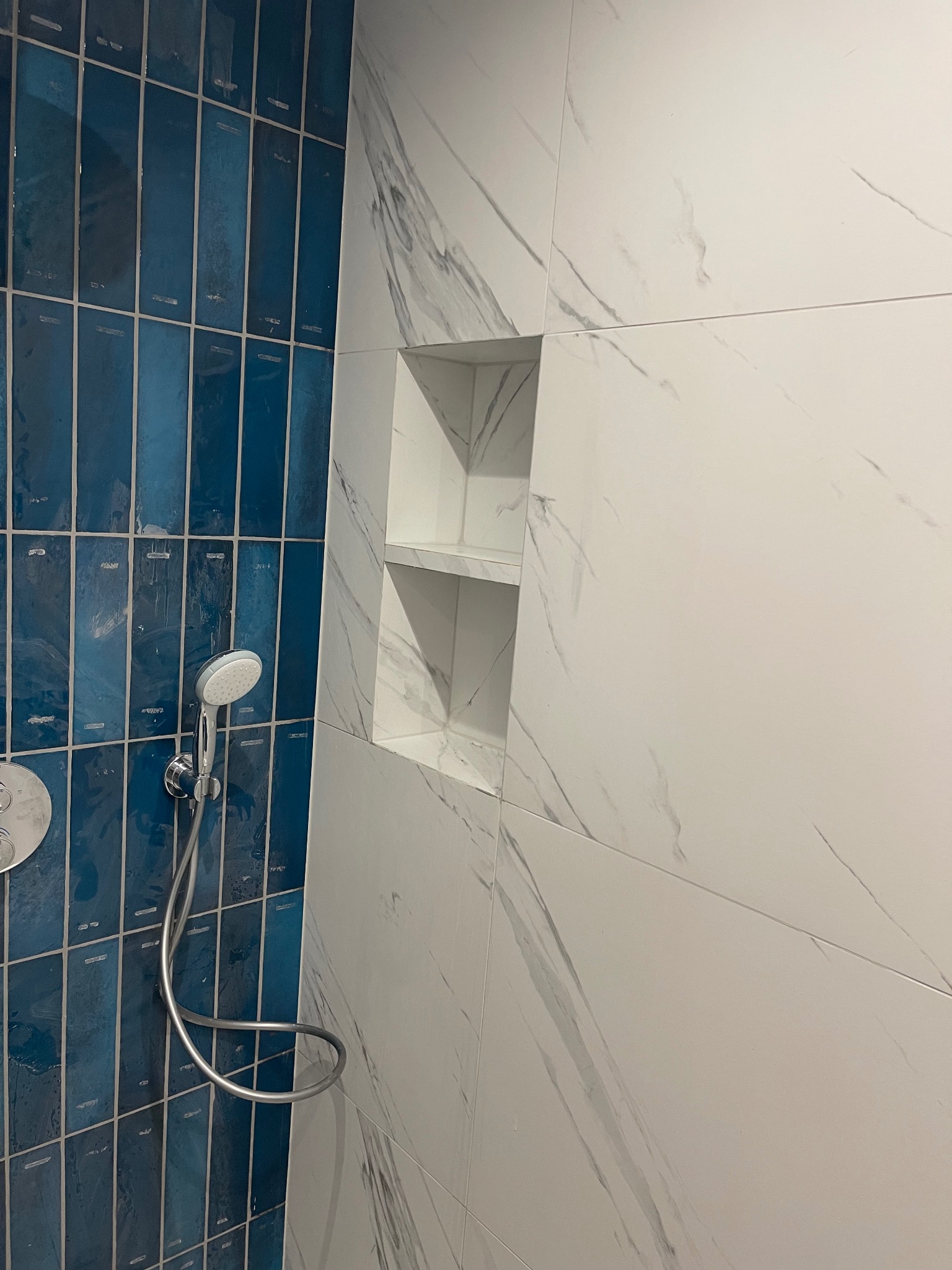 Niebiesko-biała łazienka z kabiną prysznicową, wykończona płytkami w dwóch odcieniach i wnęką. Nowoczesny design z chromowaną słuchawką prysznicową.