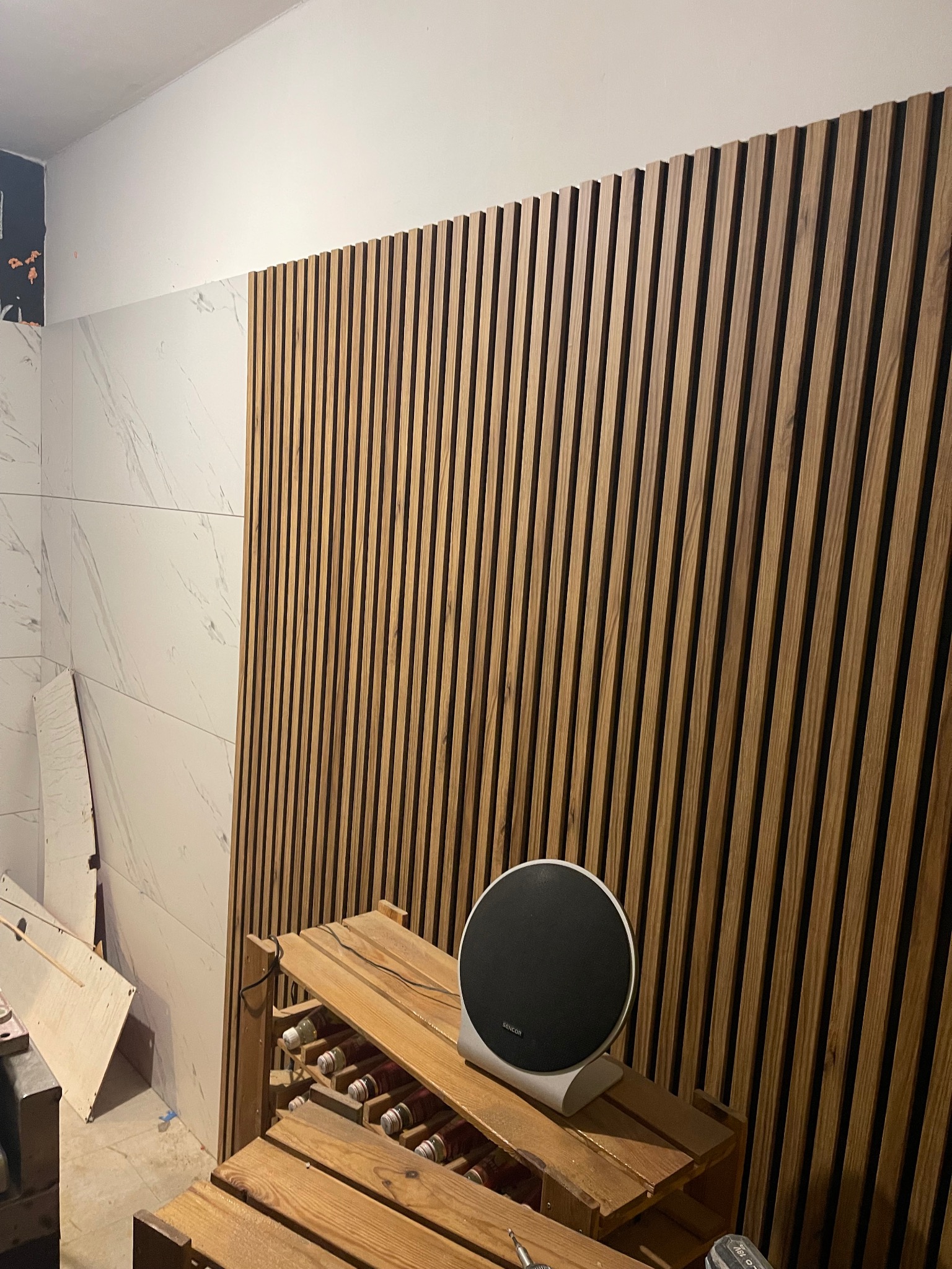 Drewniane lamele na ścianie w trakcie montażu, widoczne narzędzia i fragment ściany z marmurowymi płytkami. Częściowo wykończone wnętrze.