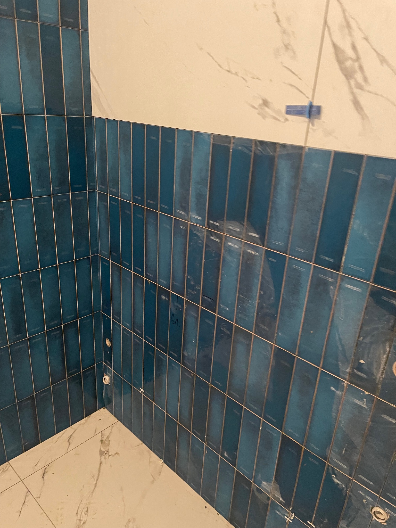 Niebieskie, pionowe płytki w połowie ściany łazienki, białe płytki na podłodze, widoczne krzyżyki dystansowe, etap prac wykończeniowych.