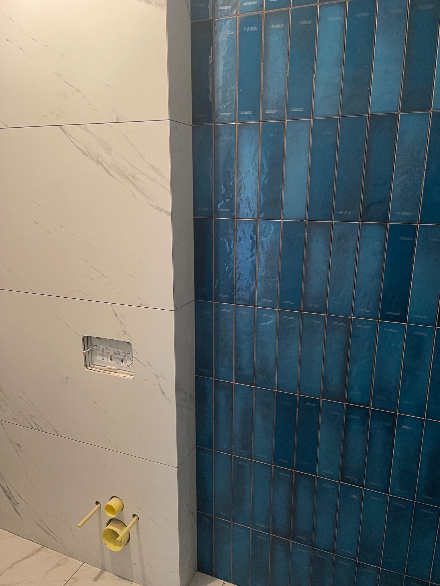 Ściana wyłożona białymi płytami imitującymi marmur i pionowymi, błyszczącymi, niebieskimi płytkami. Widoczne żółte rury i otwór na stelaż podtynkowy.