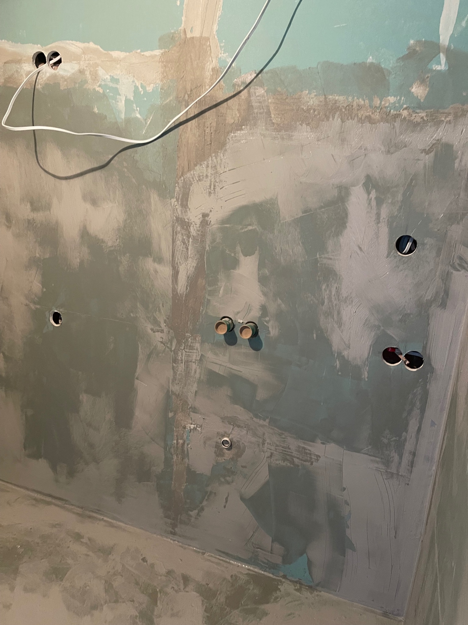 Ściana w trakcie szpachlowania z widocznymi otworami na gniazdka elektryczne i wystającymi przewodami. Surowy stan wykończenia, przygotowanie do dalszych prac.