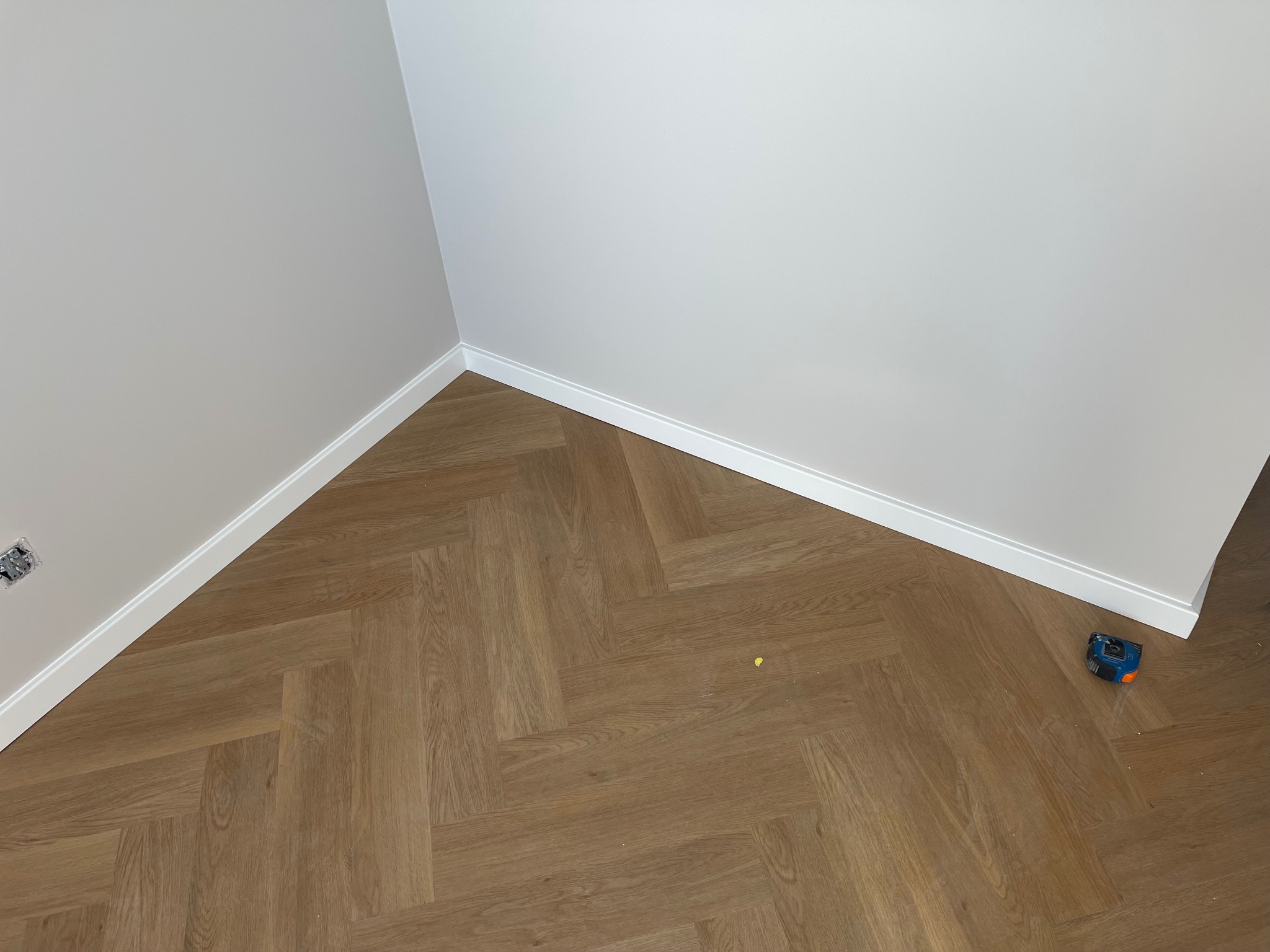 Ukończona podłoga z parkietu ułożonego w jodełkę, białe listwy przypodłogowe, ściana pomalowana na jasny kolor, widoczna puszka elektryczna i miarka.