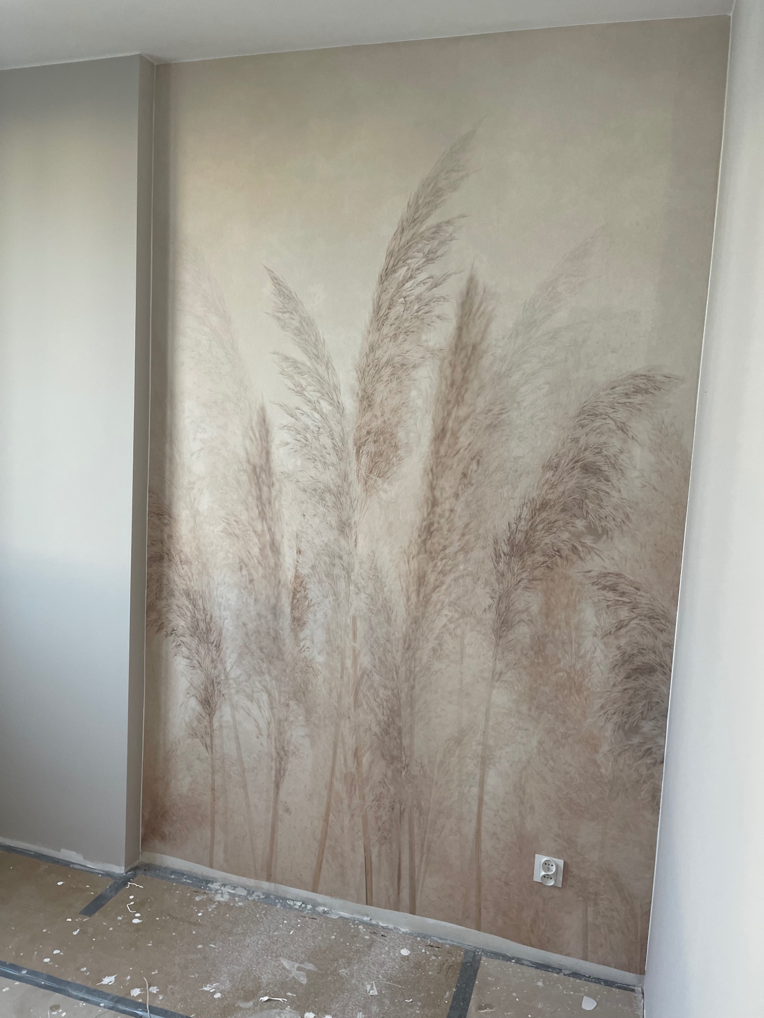 Ściana z dekoracyjną tapetą w odcieniach beżu, przedstawiającą motyw trawy pampasowej. Widoczny fragment podłogi w trakcie prac wykończeniowych i gniazdko elektryczne.
