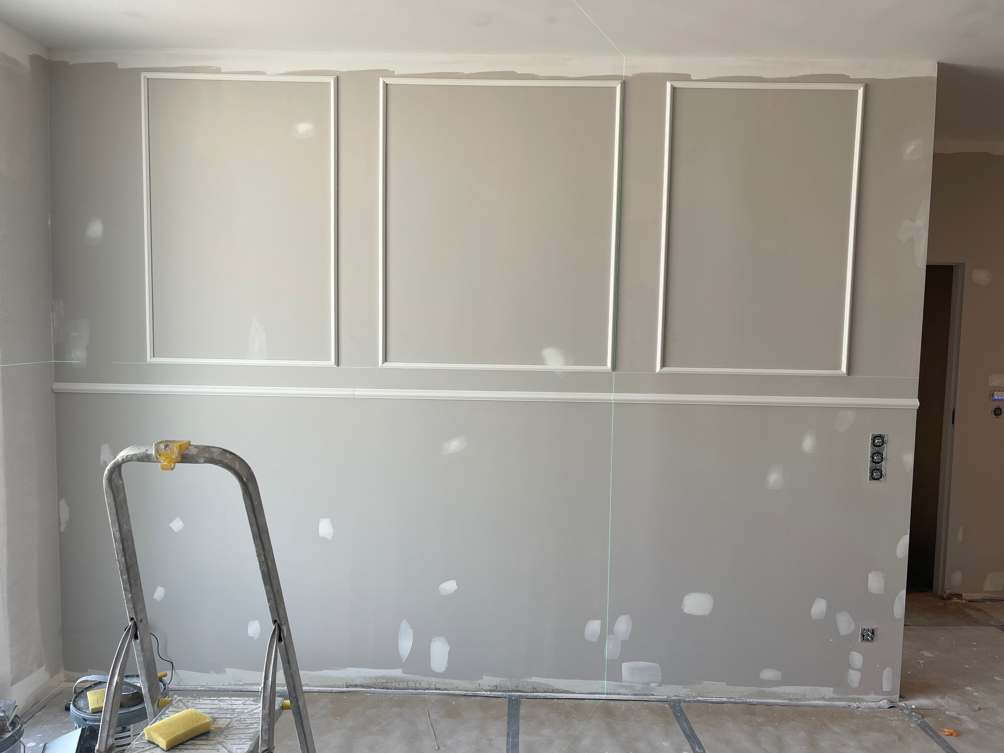 Ściana z dekoracyjnymi panelami w trakcie montażu, widoczne ślady szpachlowania i drabina malarska. Laserowy wskaźnik poziomu. Wnętrze w trakcie wykańczania.