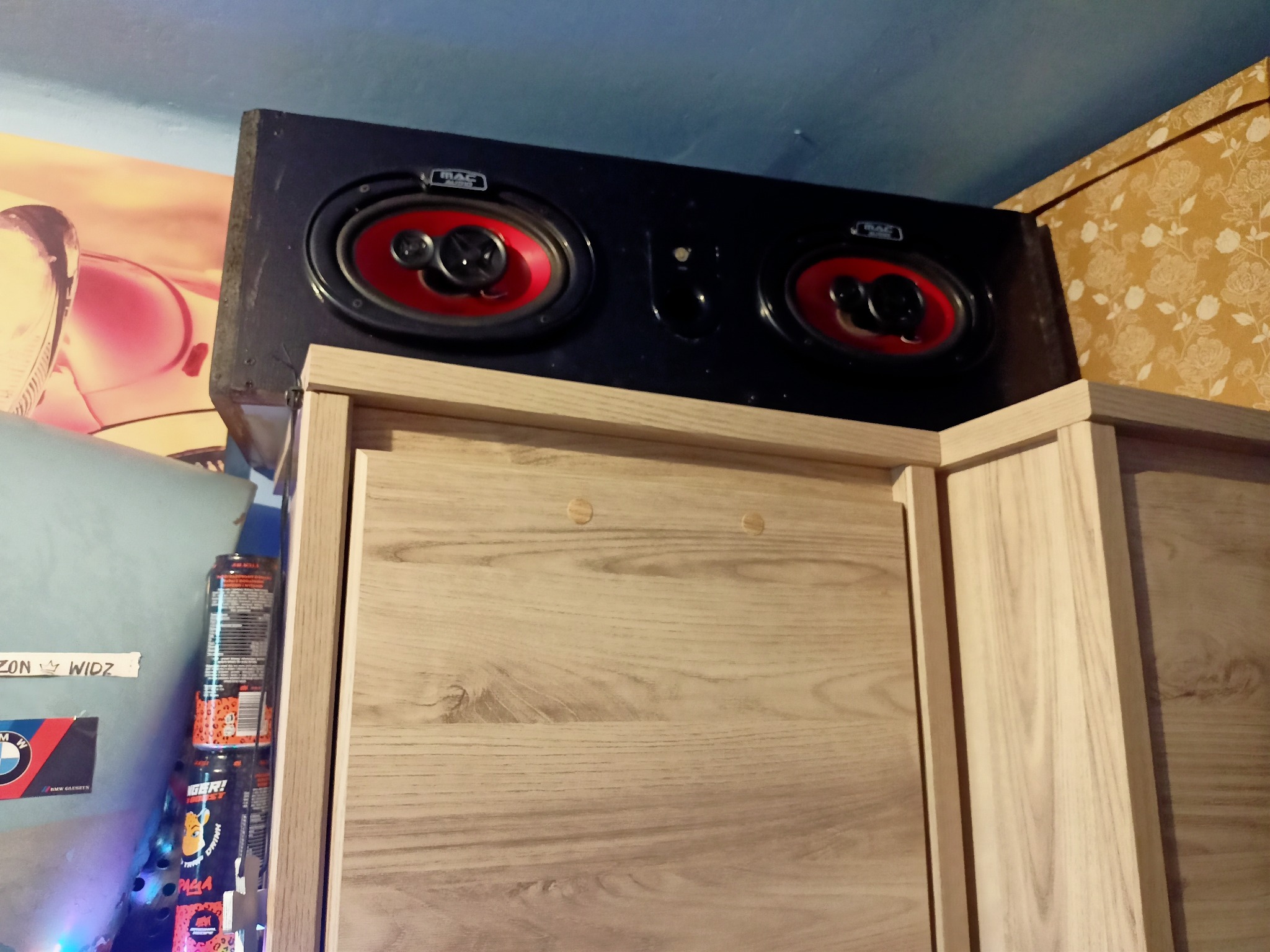 Czarna kolumna głośnikowa MAC Audio z czerwonymi membranami, umieszczona na drewnianej szafce. Widoczne puszki z napojami i naklejki na ścianie w tle.
