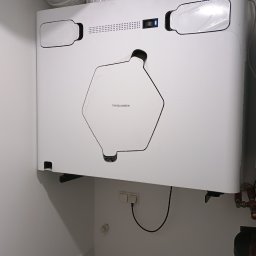 Revive - Biała jednostka rekuperacji Thessla Green zamontowana na ścianie, widoczne przewody i elementy instalacji. Minimalistyczny design, nowoczesne rozwiązanie wentylacyjne.