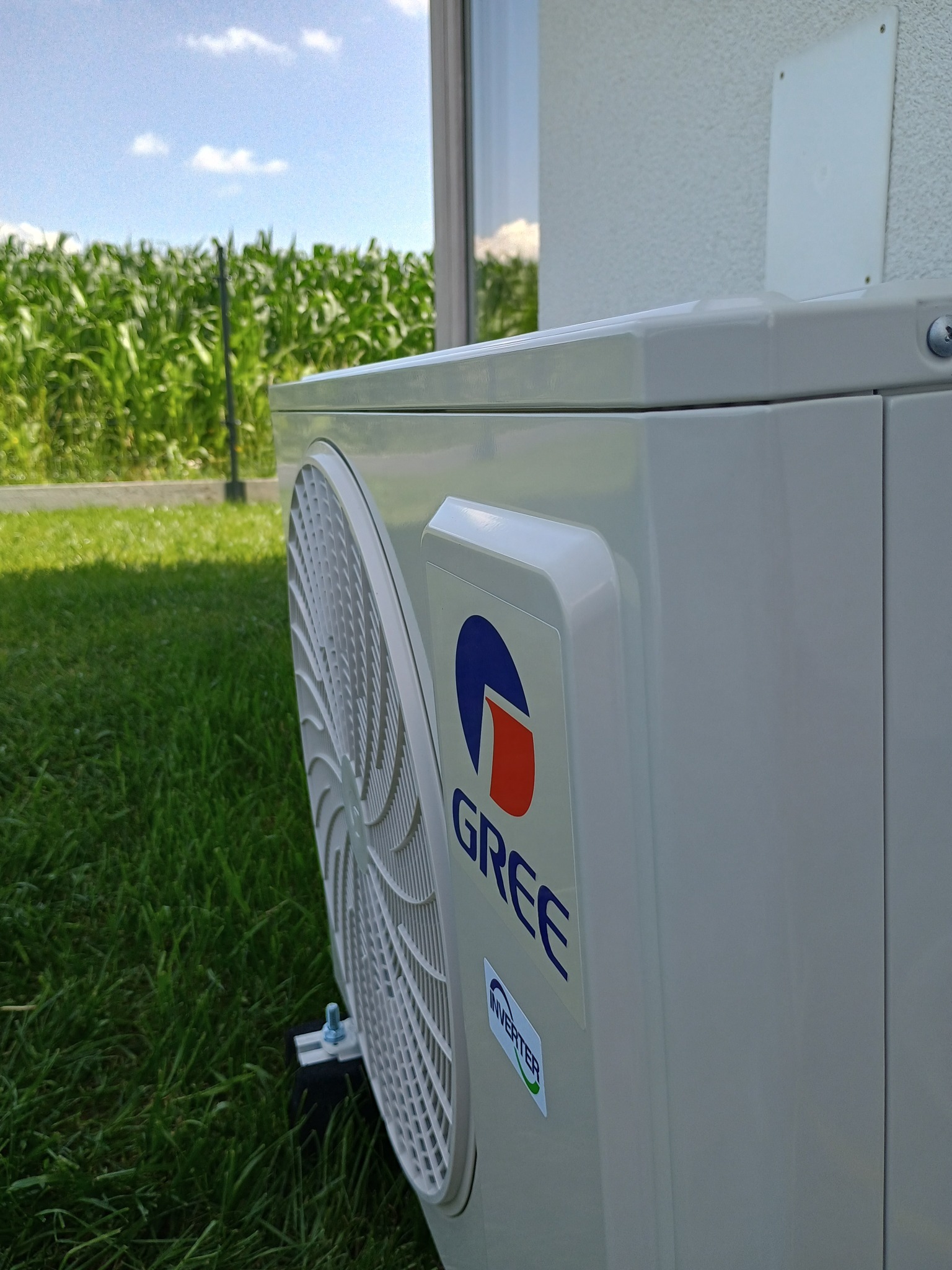 Zewnętrzna jednostka klimatyzacji GREE na trawie, widok z boku. W tle zielone pole i błękitne niebo. Widoczny fragment elewacji budynku.