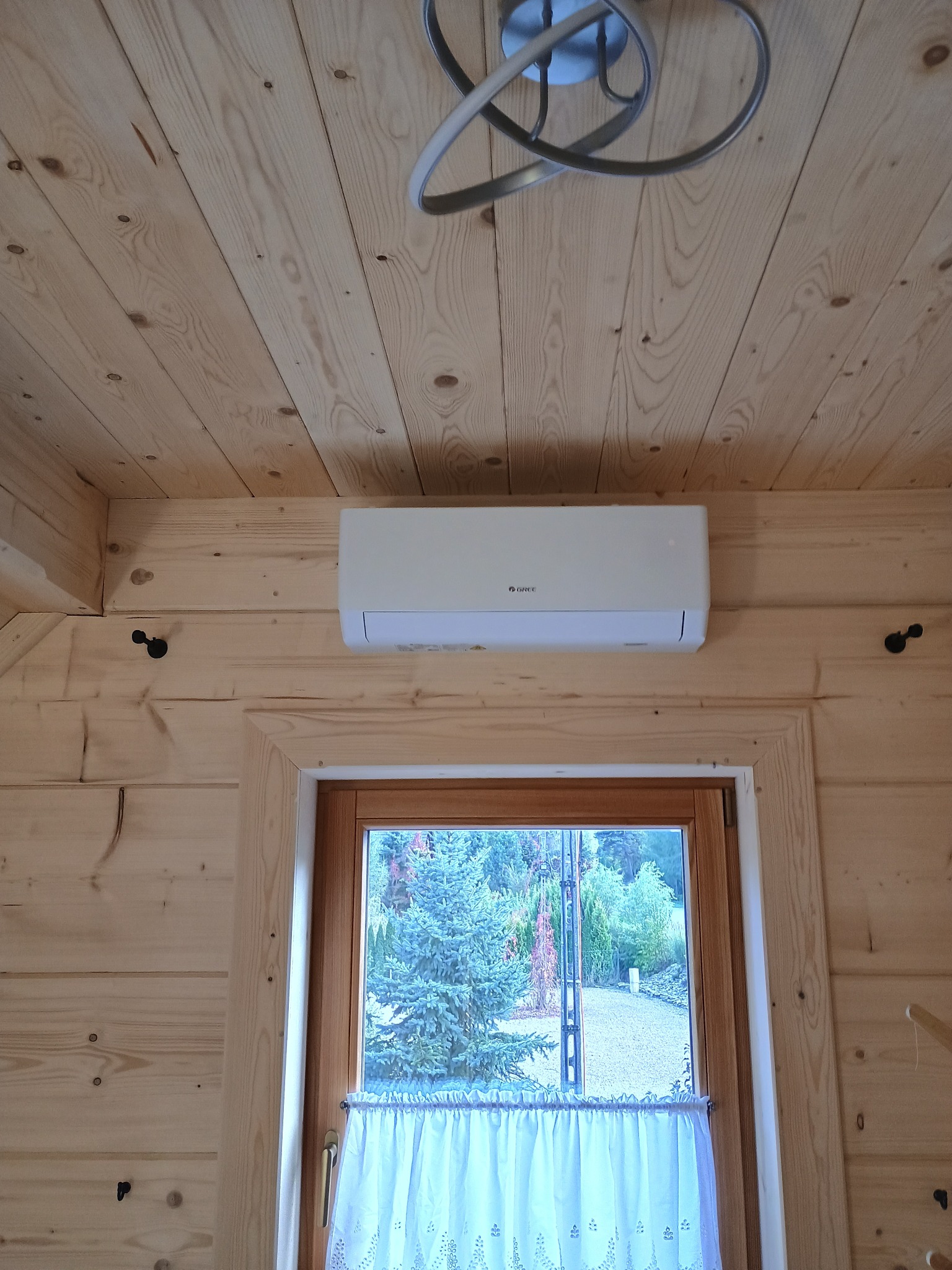 Klimatyzator Gree zamontowany w drewnianym domu w Biertowicach. Widok na okno z firanką i zielony ogród. Nowoczesna lampa sufitowa.
