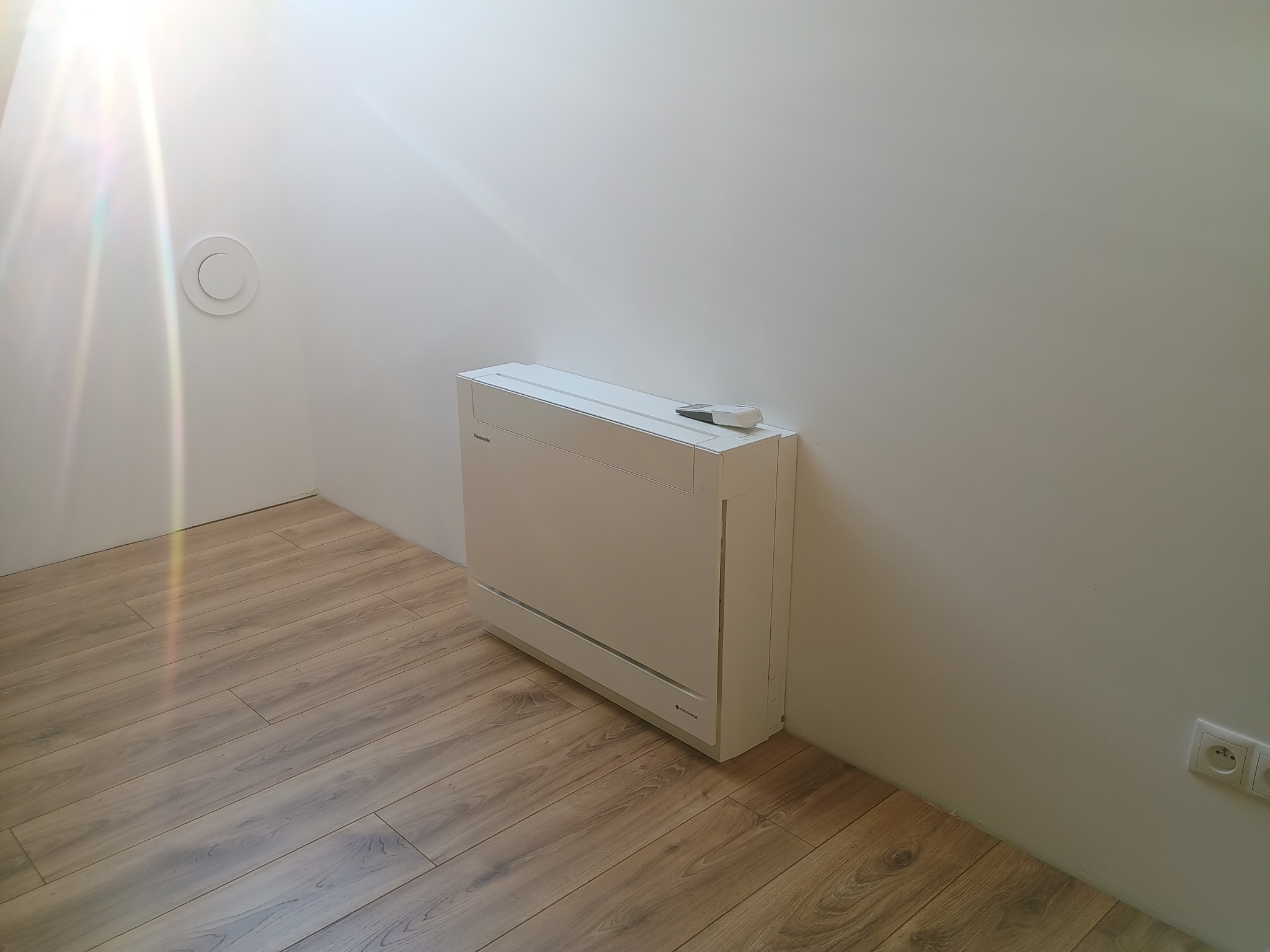 Biały klimatyzator podłogowy Panasonic w minimalistycznym wnętrzu z drewnianą podłogą i białymi ścianami. Widoczny pilot na urządzeniu i okrągły nawiewnik sufitowy.