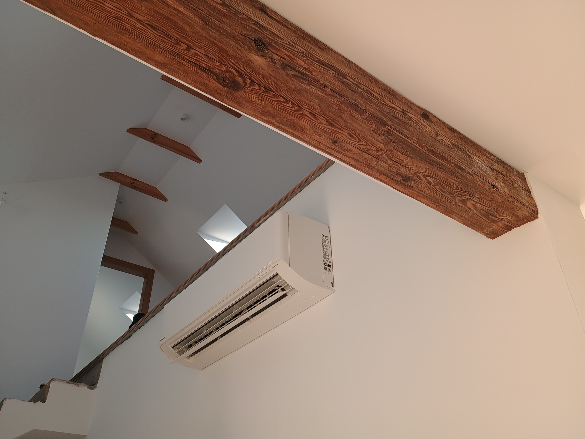 Biały klimatyzator ścienny zamontowany w jasnym wnętrzu z drewnianymi belkami stropowymi, widok z dołu, ukośny kadr.