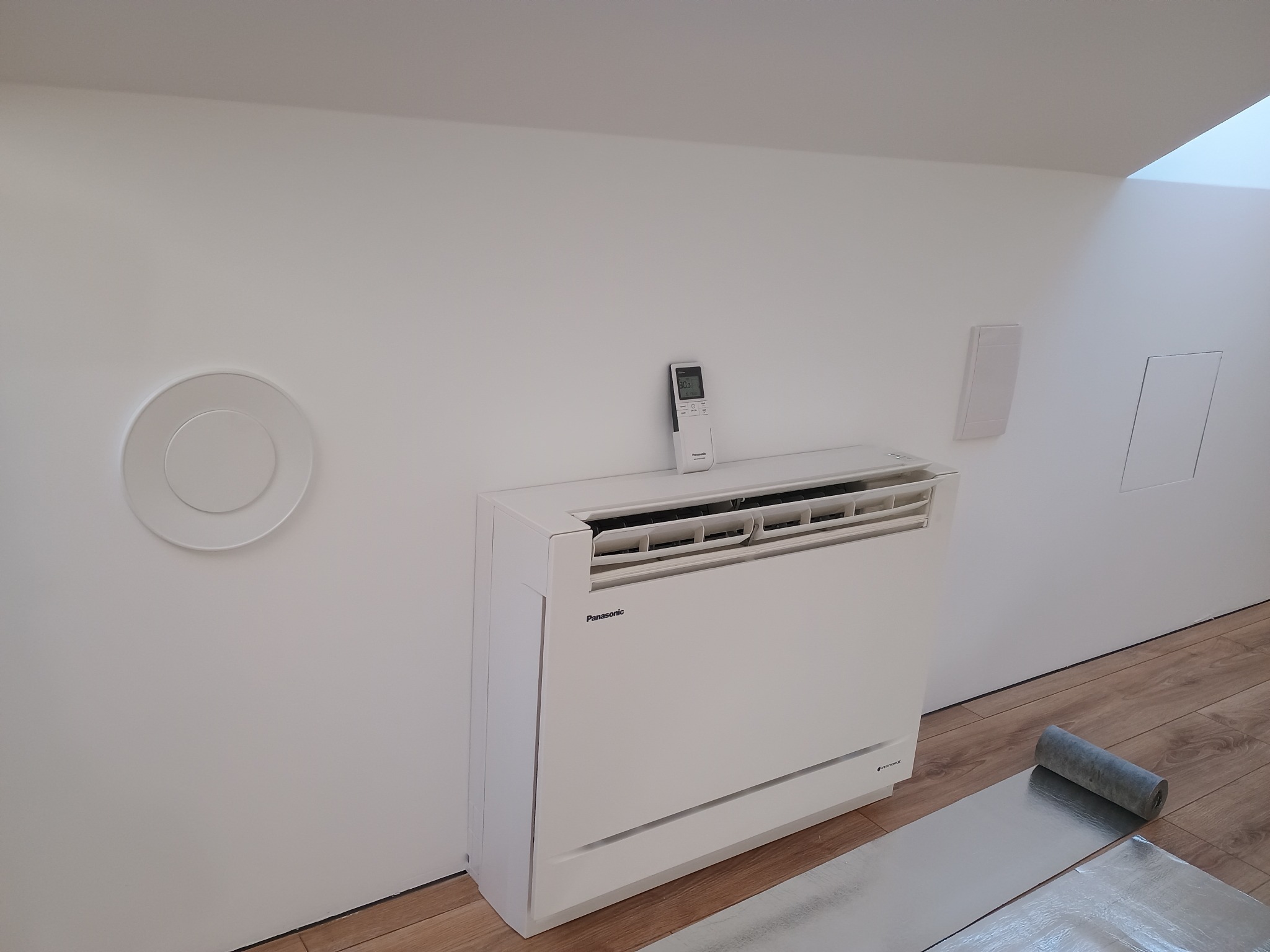 Biały klimatyzator podłogowy Panasonic w jasnym wnętrzu ze skośnym sufitem i drewnianą podłogą. Na ścianie okrągły nawiewnik i pilot na urządzeniu.