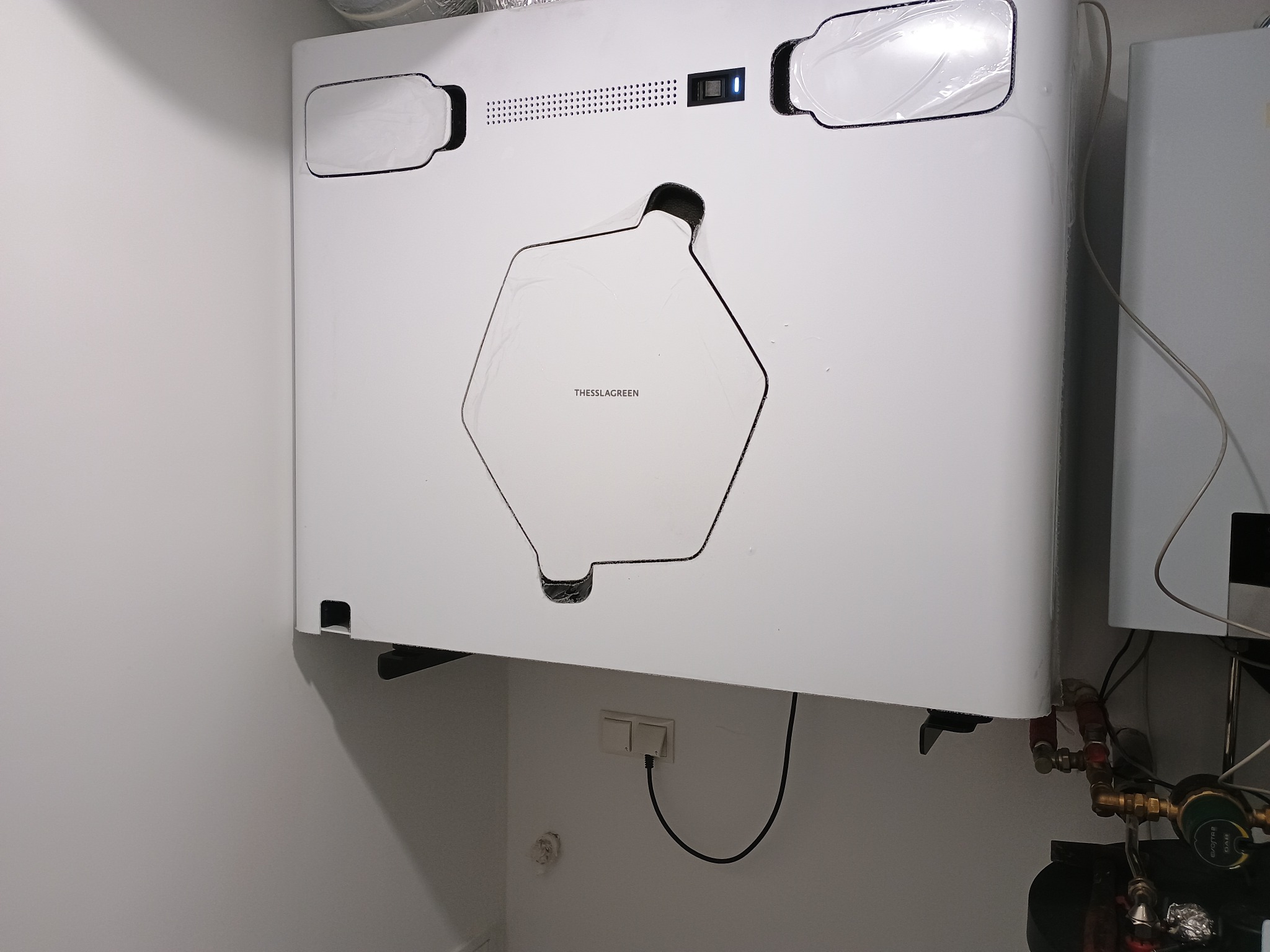 Biała jednostka rekuperacji Thessla Green zamontowana na ścianie, widoczne przewody i elementy instalacji. Minimalistyczny design, nowoczesne rozwiązanie wentylacyjne.