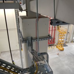 PRZEMYSŁAW KIJEWSKI SOLUTION - Metalowe korytka kablowe zamontowane na słupie w hali przemysłowej, widoczne kable wewnątrz. W tle podnośnik koszowy i instalacja przeciwpożarowa.