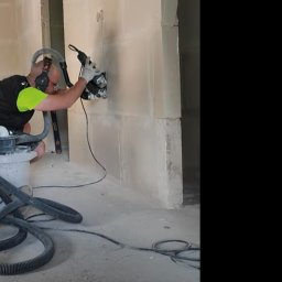 PRZEMYSŁAW KIJEWSKI SOLUTION - Pracownik w słuchawkach tnie ścianę z betonu komórkowego szlifierką kątową z odkurzaczem przemysłowym. Widoczne przewody i poziomica.