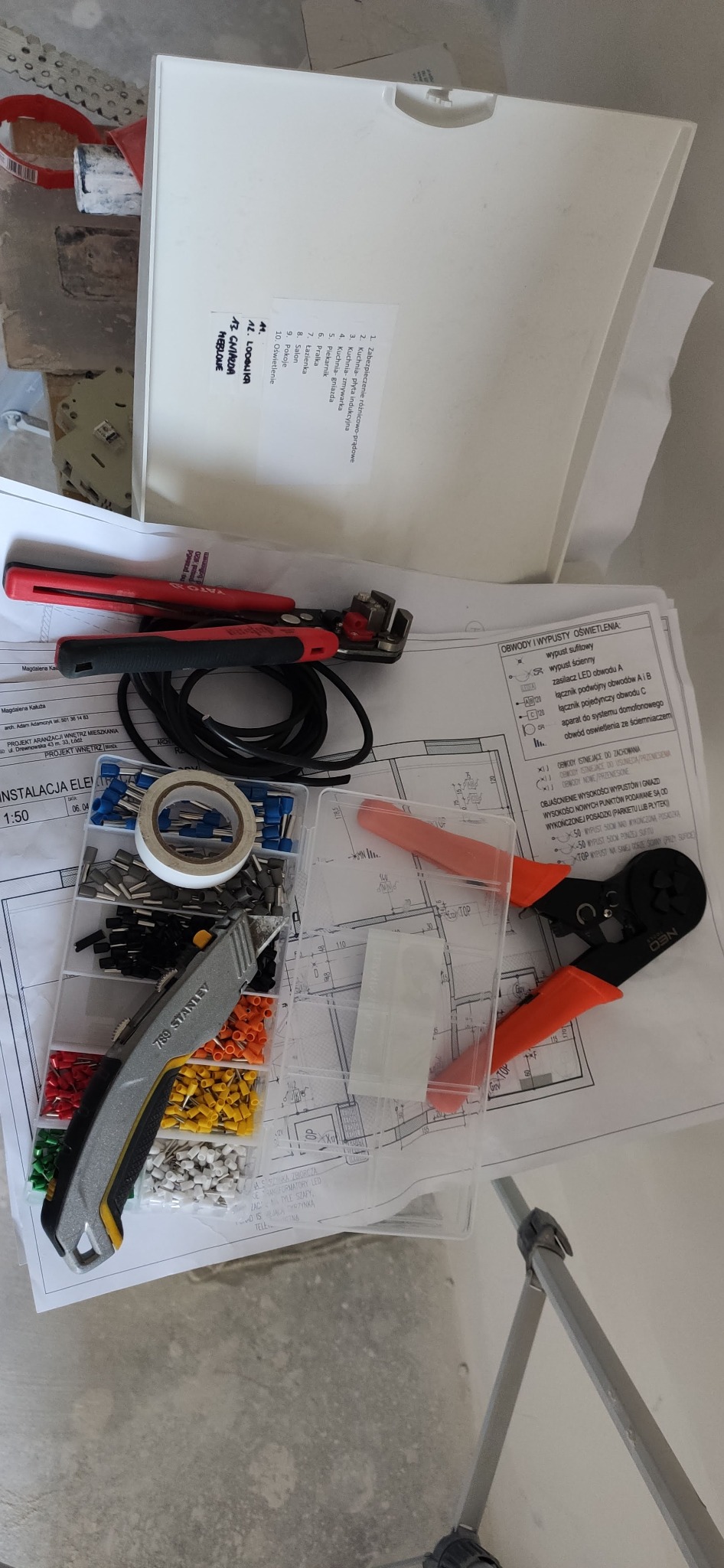 Plan instalacji elektrycznej z narzędziami: nóż, zaciskarka, wsuwki, taśma izolacyjna. Widoczne obwody oświetlenia i gniazda. Elementy instalacji na stole.