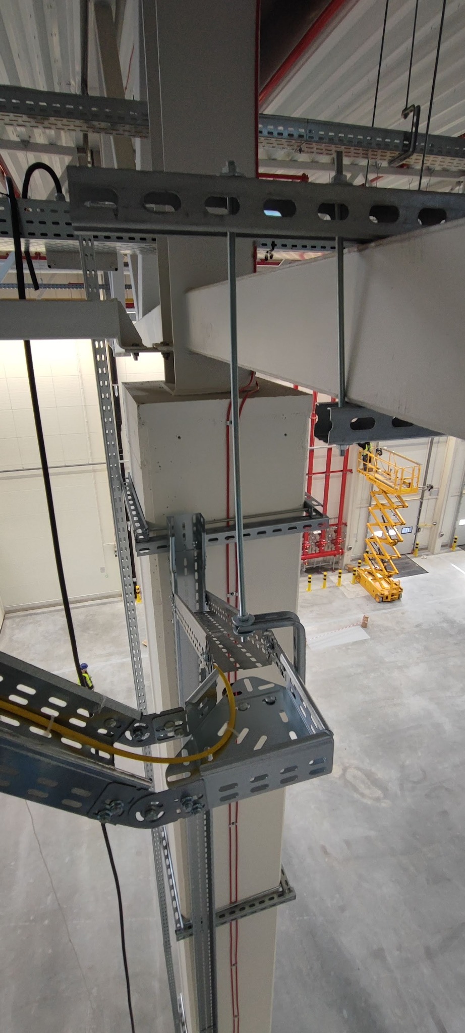 Metalowe korytka kablowe zamontowane na słupie w hali przemysłowej, widoczne kable wewnątrz. W tle podnośnik koszowy i instalacja przeciwpożarowa.