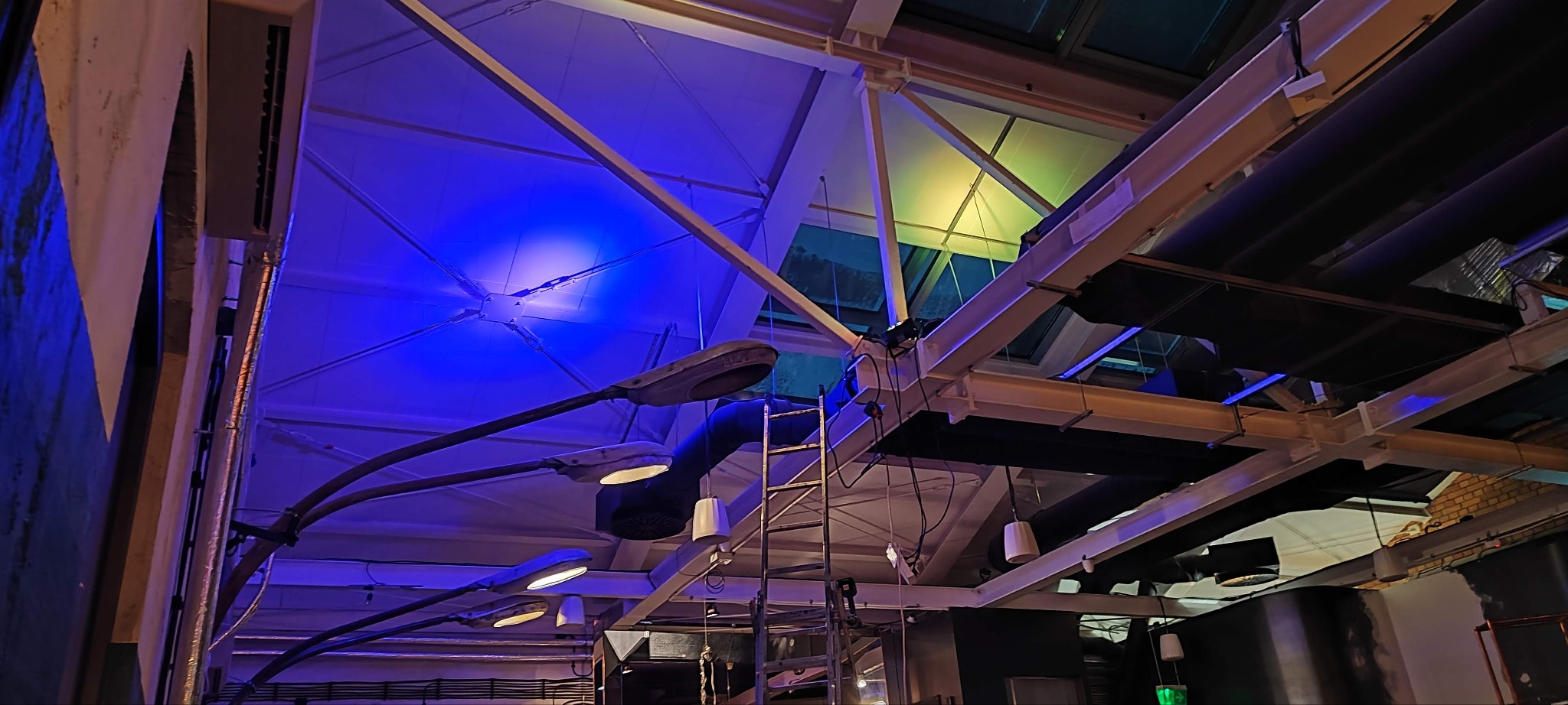 Instalacja oświetlenia przemysłowego: lampy uliczne wewnątrz hali, widoczne elementy konstrukcyjne dachu i drabina. Oświetlenie w odcieniach niebieskiego i żółtego.