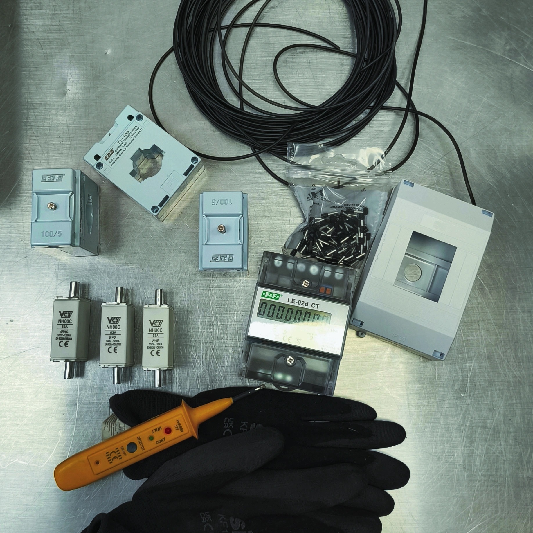 Elementy instalacji elektrycznej: bezpieczniki, miernik, transformatory prądowe, przewody i tester napięcia na tle roboczych rękawic i metalowego blatu.