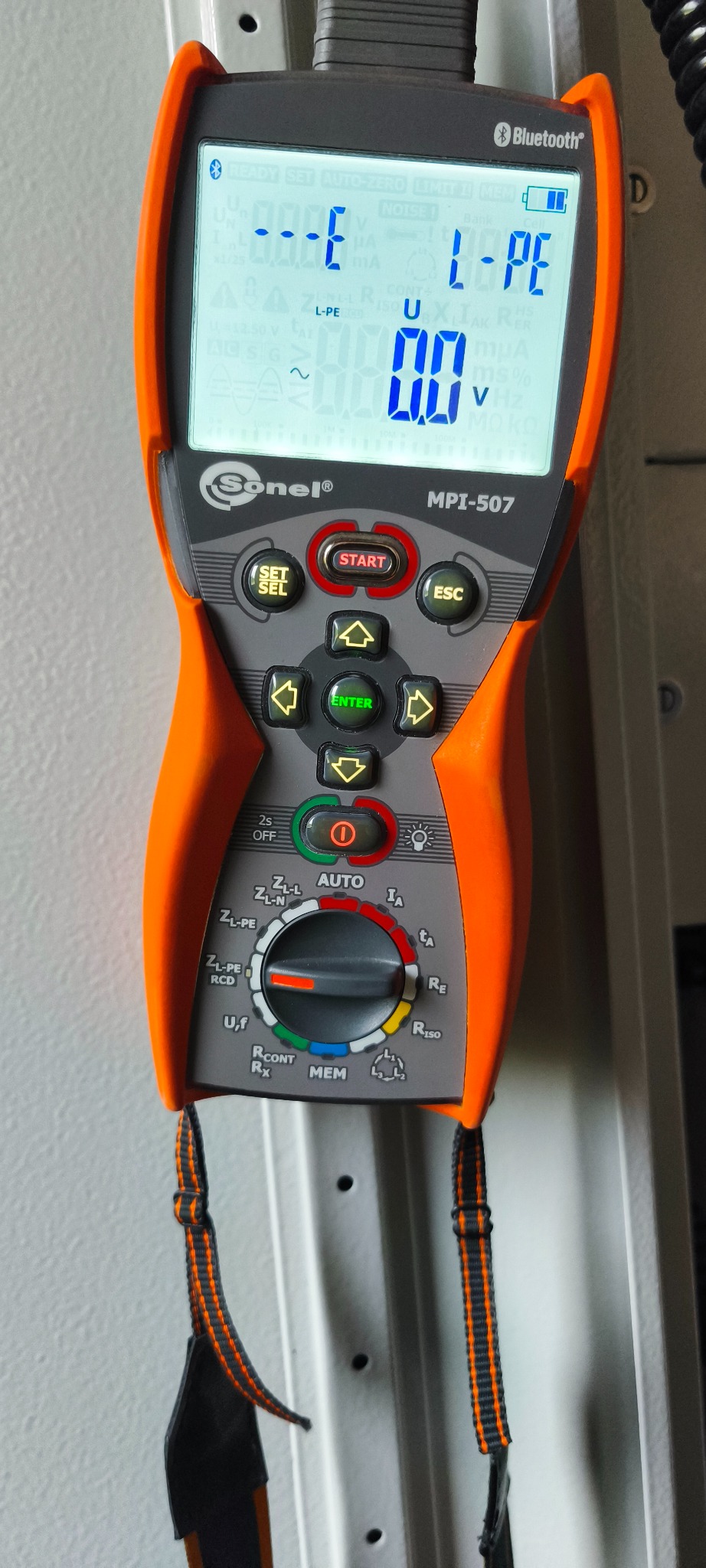 Miernik Sonel MPI-507 z wyświetlaczem wskazującym pomiar L-PE, zawieszony na metalowej szafce elektrycznej. Widoczne przyciski i pokrętło wyboru funkcji.