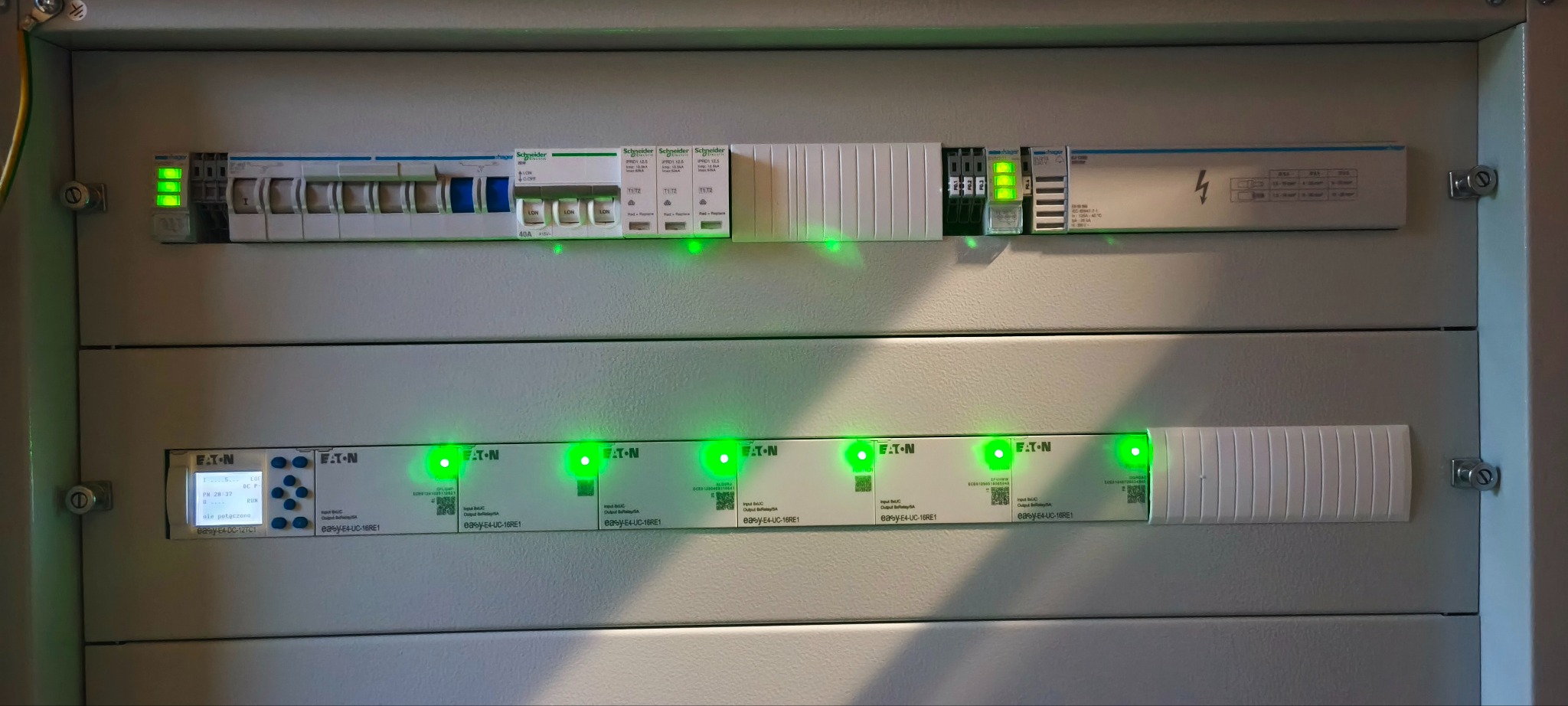 Rozdzielnica elektryczna z modułami Eaton i Schneider, z zielonymi diodami LED sygnalizującymi pracę. Widoczne zabezpieczenia i sterowniki. Szara obudowa.
