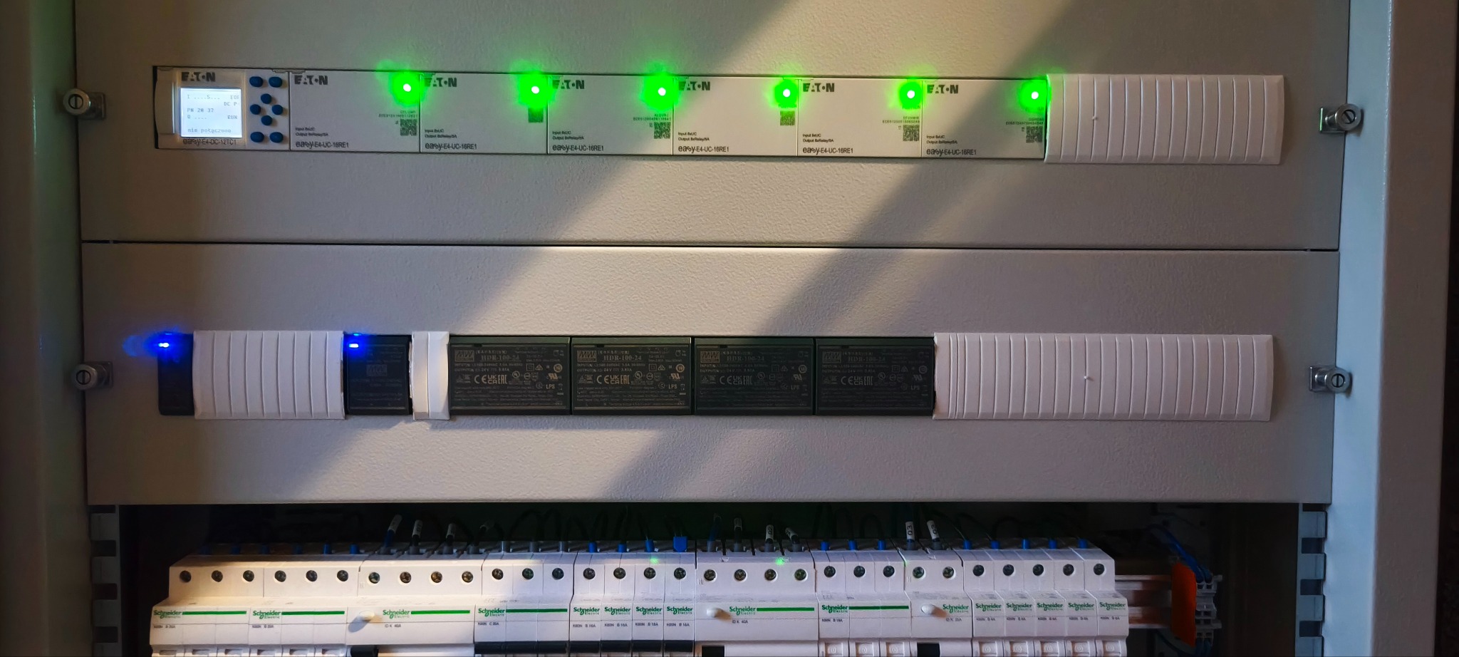 Rozdzielnica elektryczna z modułami Eaton i Schneider, z zielonymi i niebieskimi diodami LED, widok z bliska na zainstalowane zabezpieczenia i okablowanie.